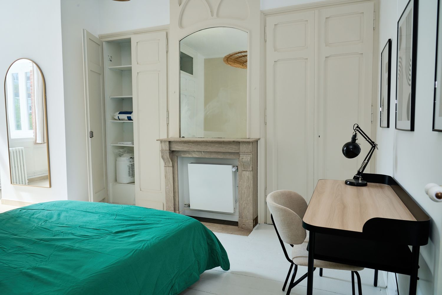 Elegant bedroom with green bedding, ornate fireplace with mirror, built-in wardrobe, and wooden desk with lamp.