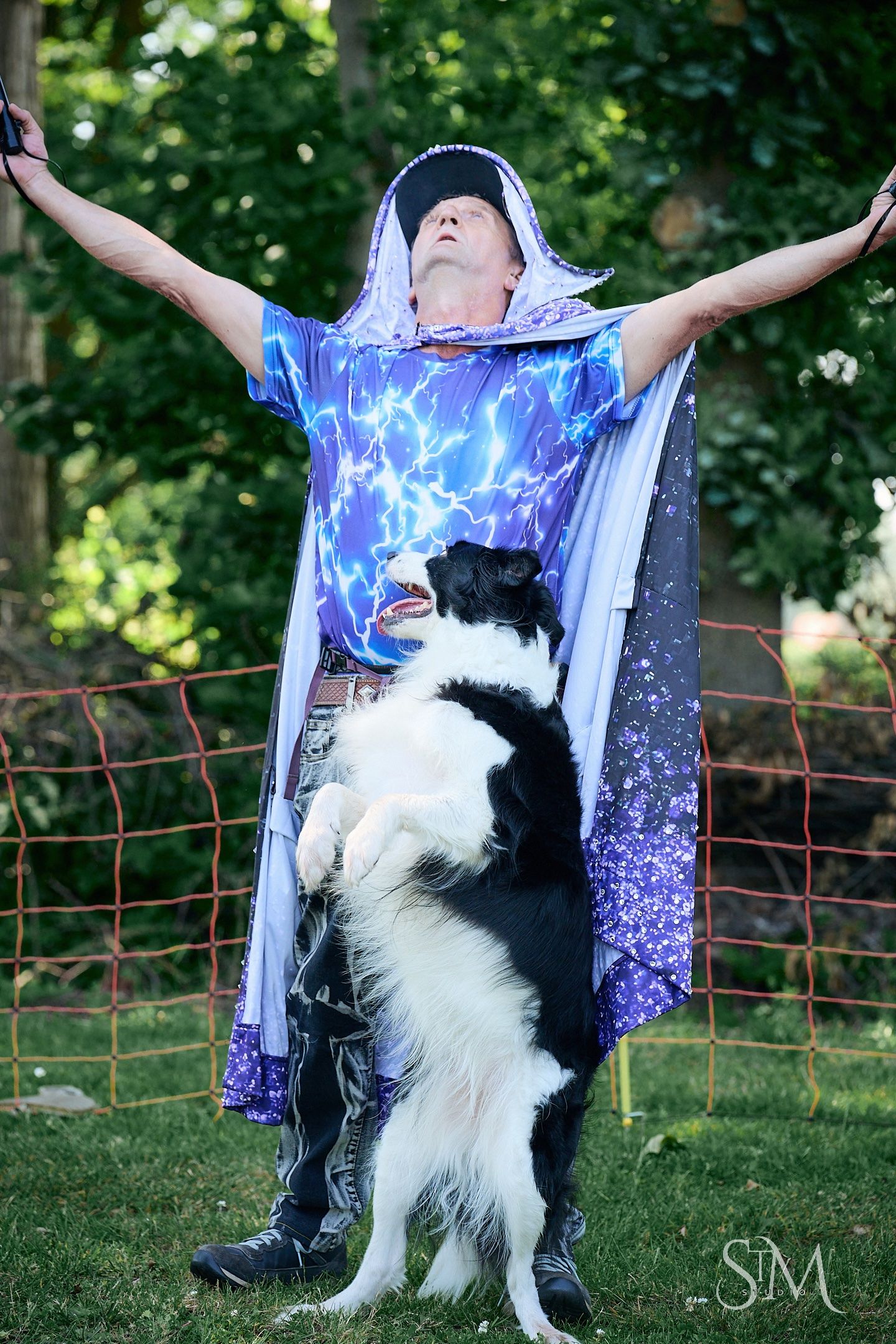 Man in lightning bolt cape with arms raised while a Border Collie stands on hind legs against him outdoors.