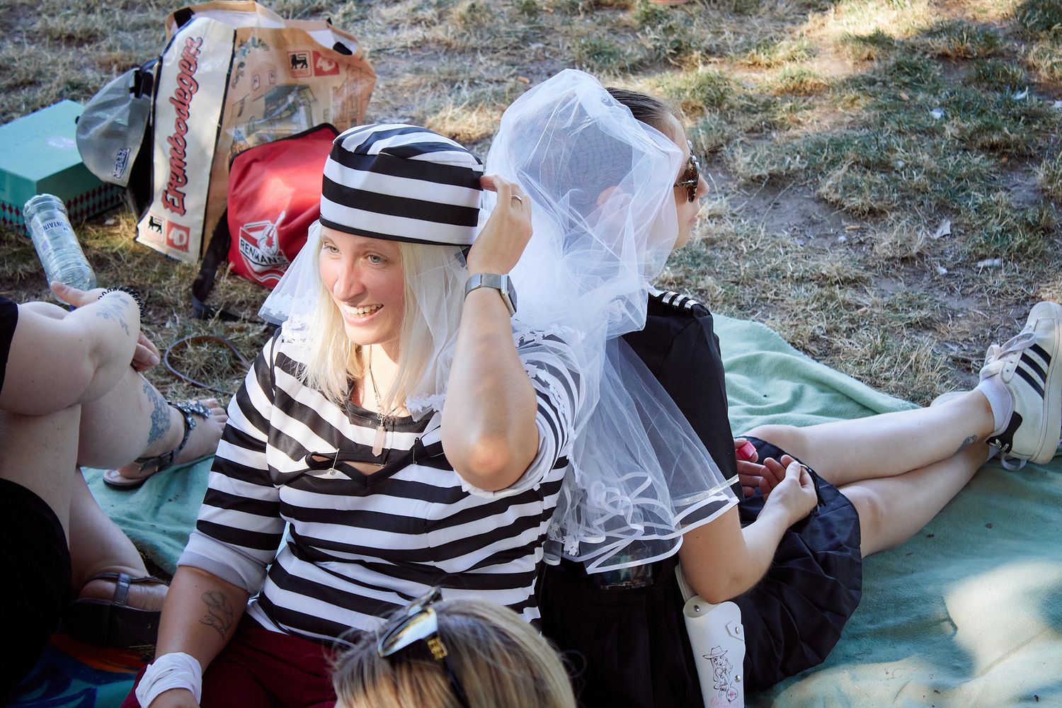 Women in striped costume and veil laughing at outdoor picnic gathering on grass blanket.