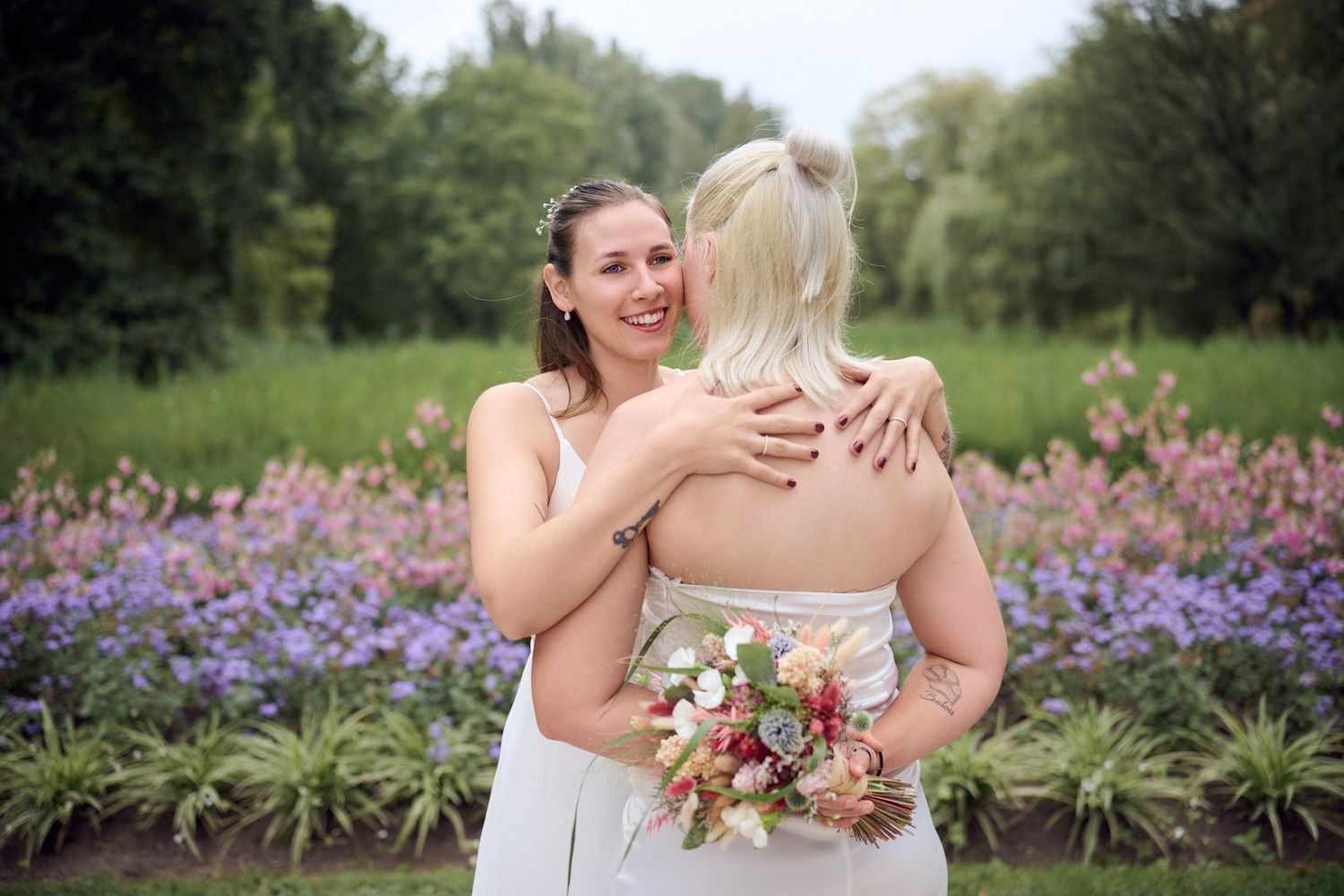 Two brides embrace joyfully in a garden with purple flowers, one holding a colorful bouquet on their wedding day.
