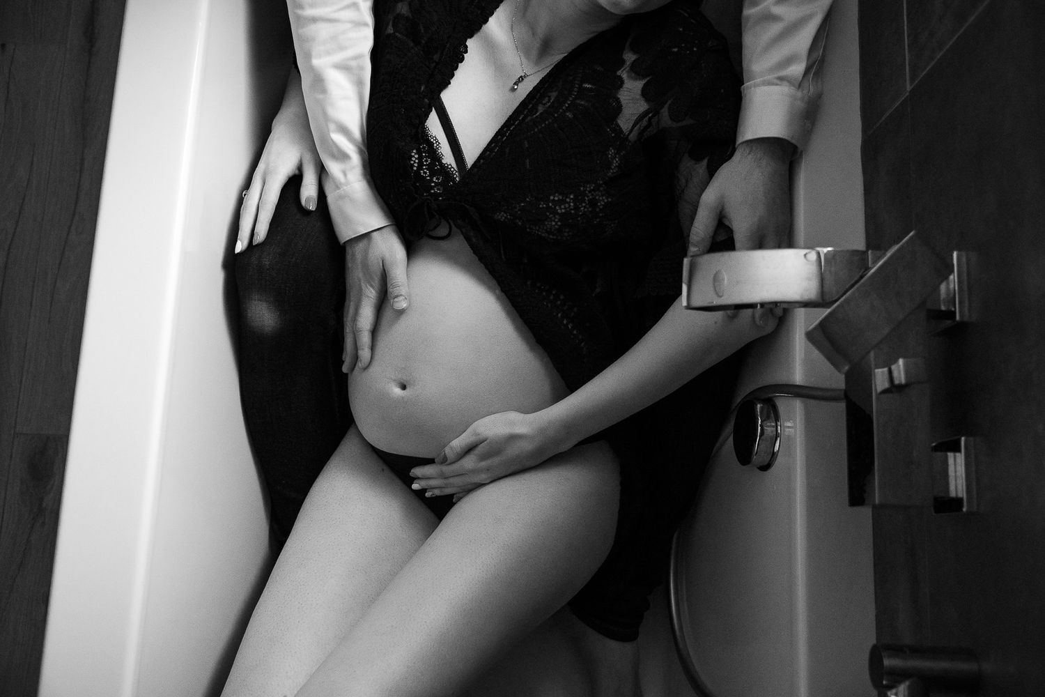Black and white photo of a pregnant woman in lace lingerie with a partner's hands gently cradling her baby bump.