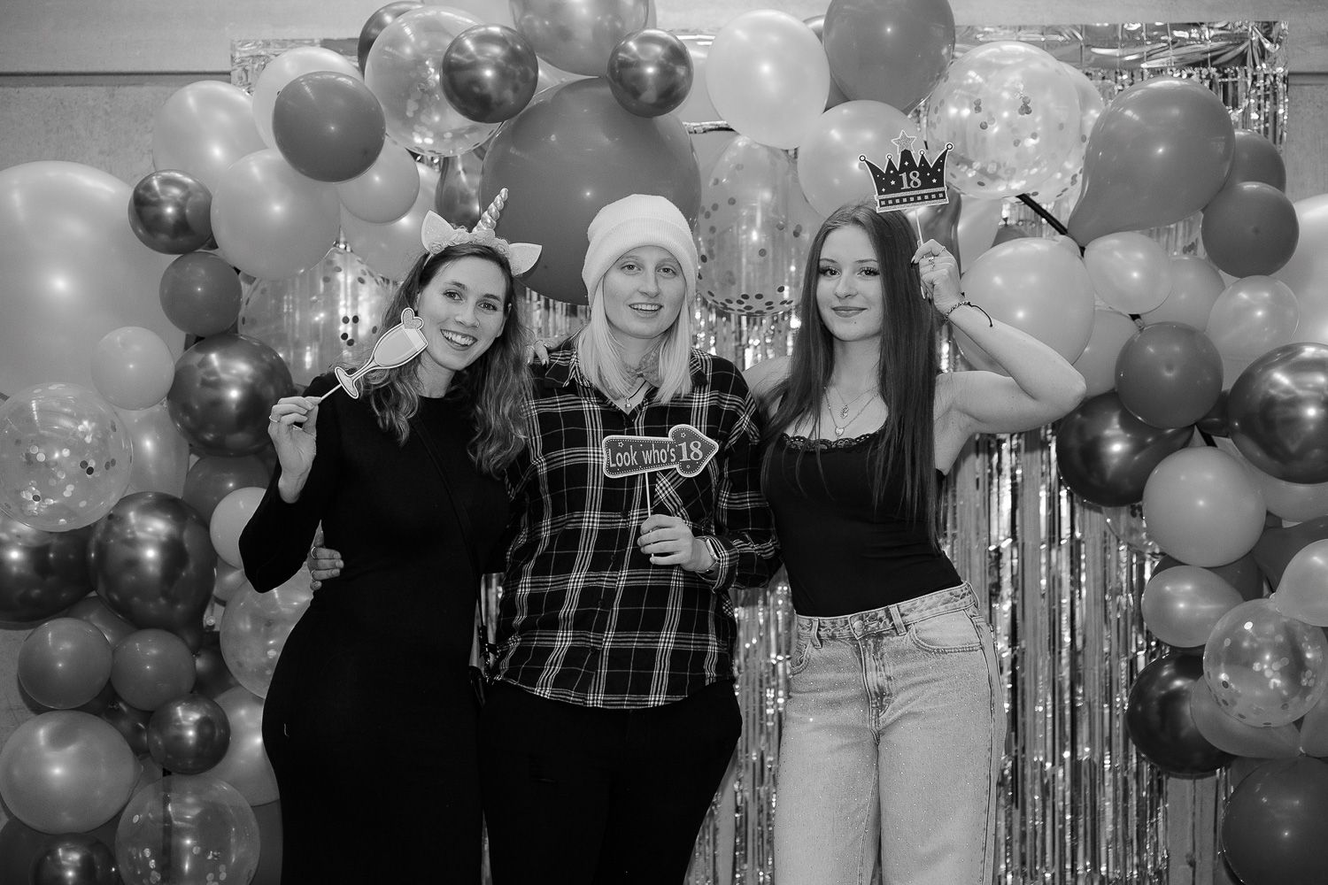 Three women pose with party props in front of a balloon arch backdrop at a festive celebration event.