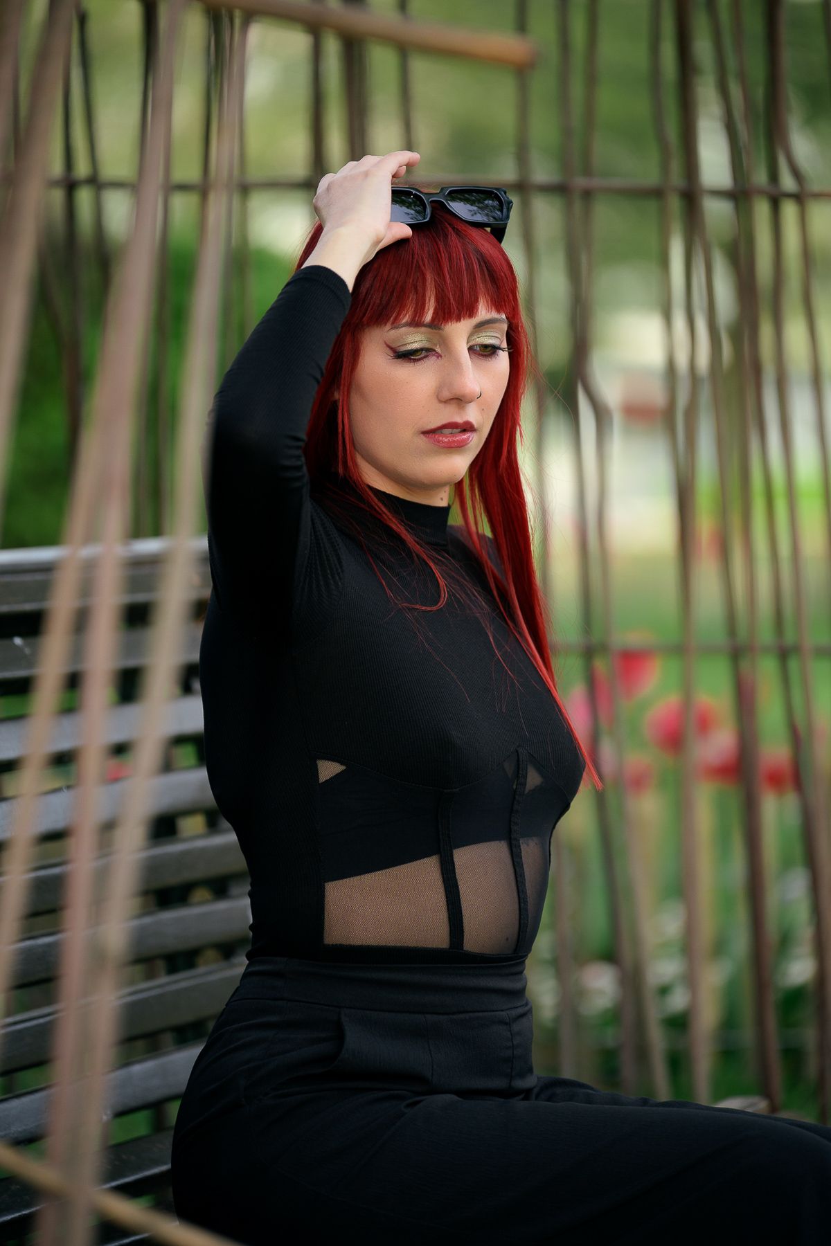Red-haired woman in black outfit sitting on bench in park, posing with sunglasses on head.