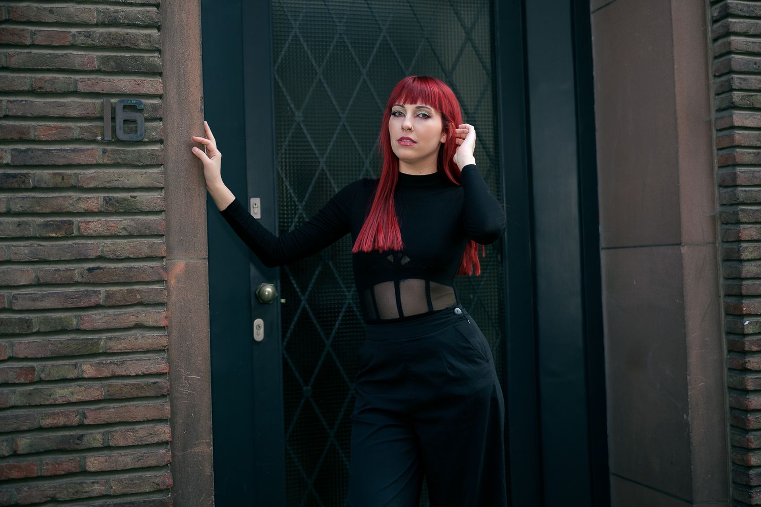 Woman with red hair in black outfit posing against a dark door on a brick building exterior.