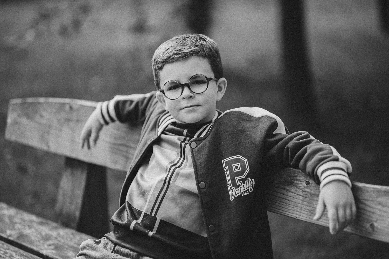 Black and white portrait of a boy in a varsity jacket relaxing on a wooden bench.