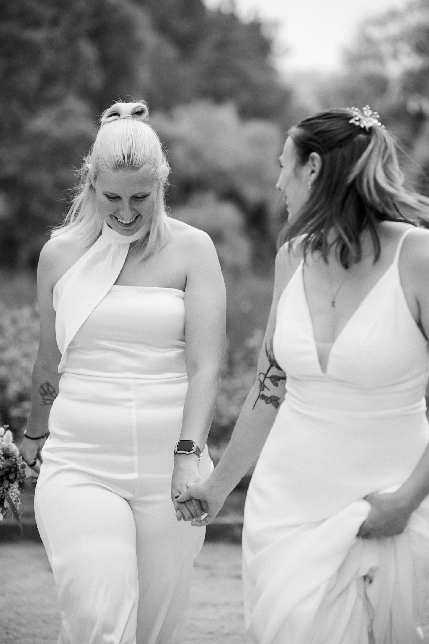 Two brides in white gowns hold hands and smile while walking outdoors in a black and white wedding photo.