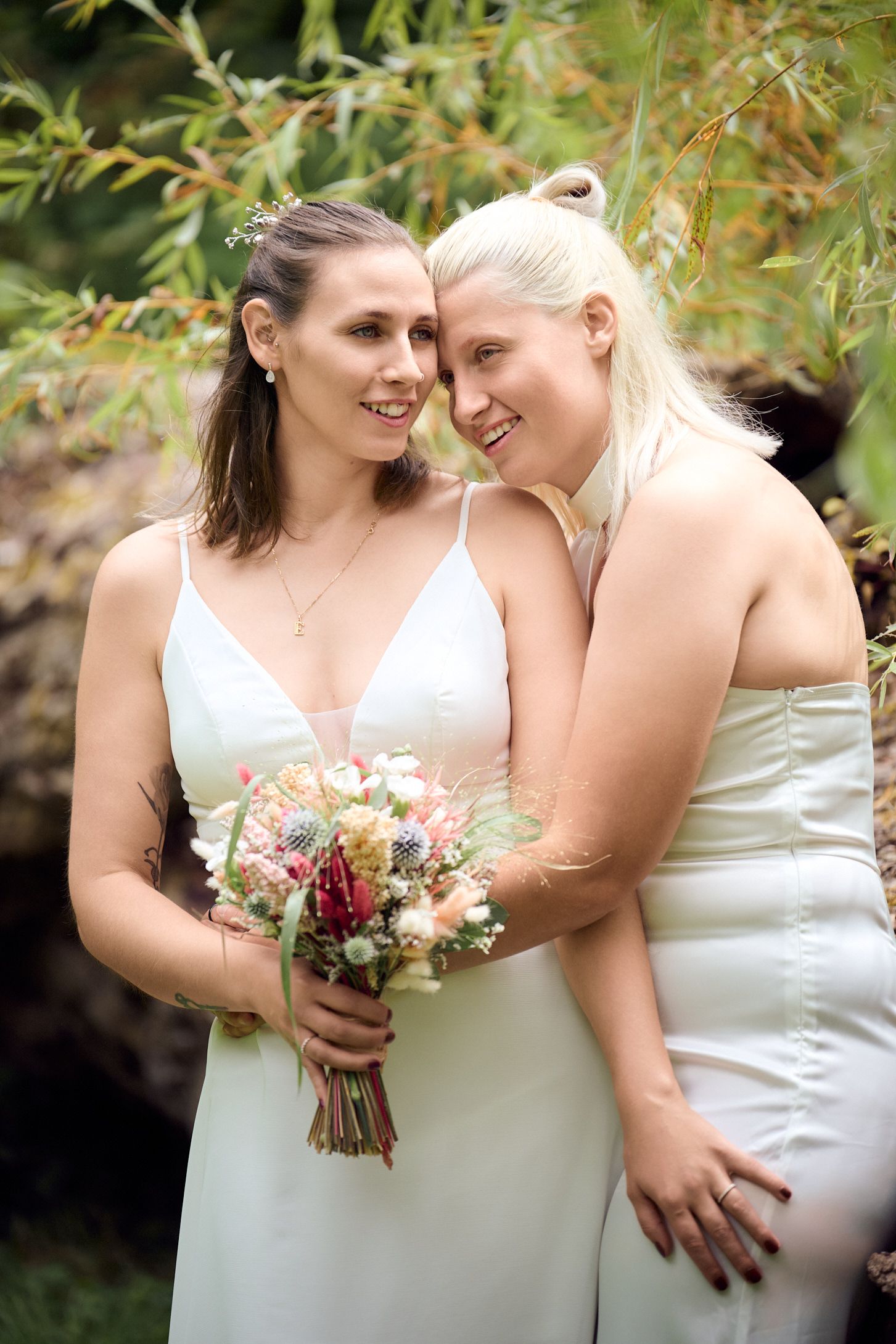 Two brides in white dresses share a tender moment, one holding a pastel bouquet, surrounded by lush greenery.