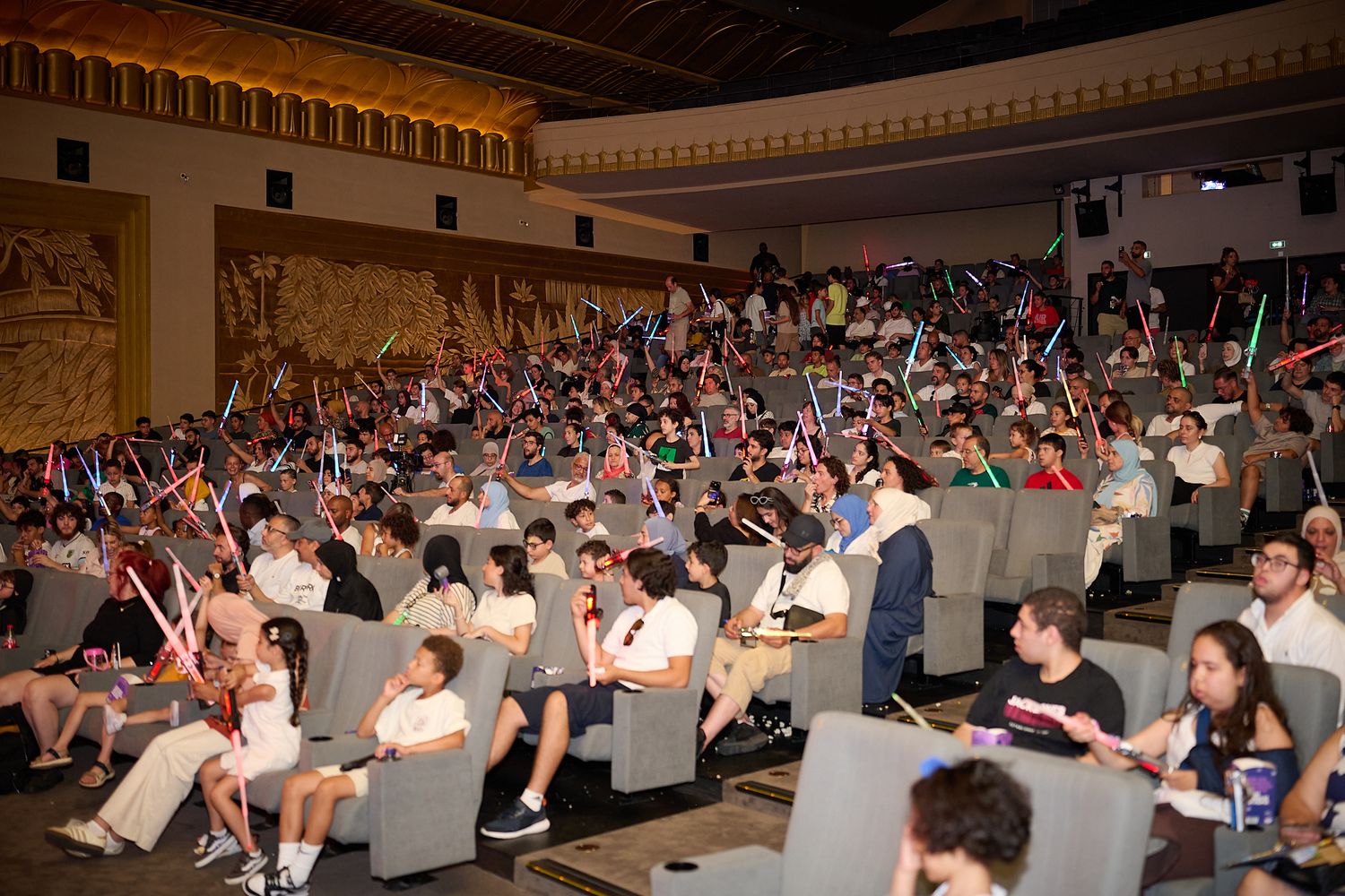 Crowd of Star Wars fans holding colorful lightsabers in a theater auditorium during a special screening event.
