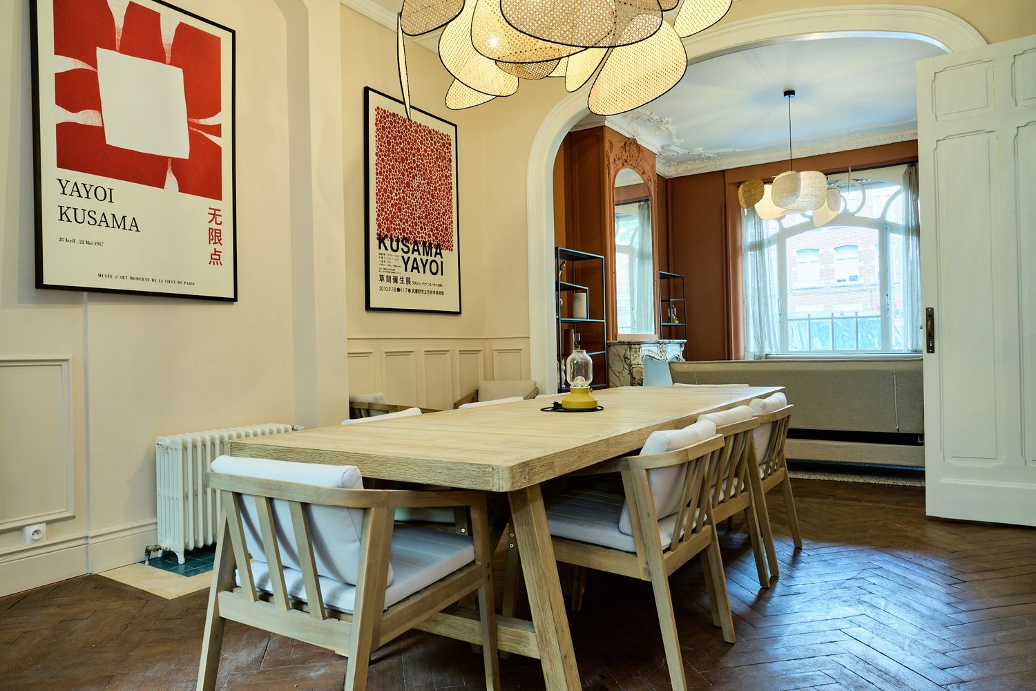 Modern dining room with wooden table, Yayoi Kusama art prints, arched bookcase, herringbone floors, and elegant pendant lighting.
