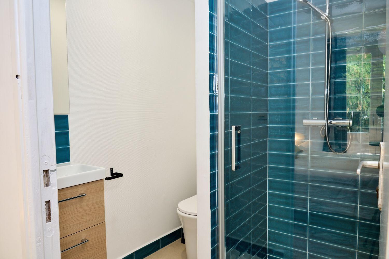 Modern bathroom with teal subway tile walk-in shower, glass door, toilet, and wooden vanity unit.