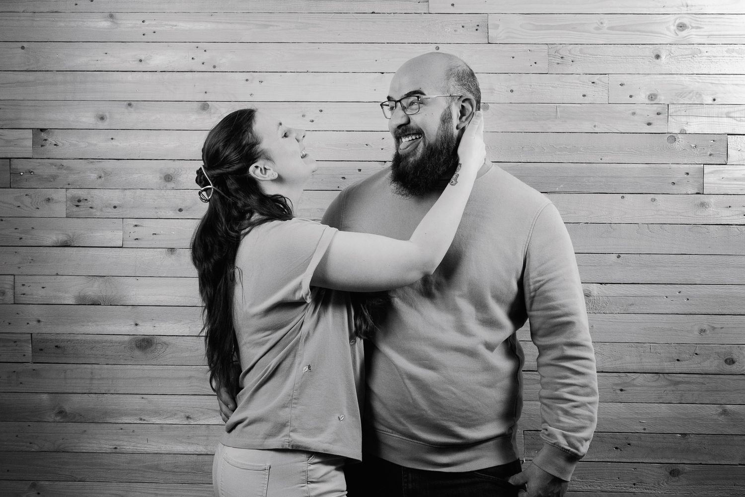 Black and white photo of a woman playfully touching a bearded bald man wearing glasses against a wood wall.