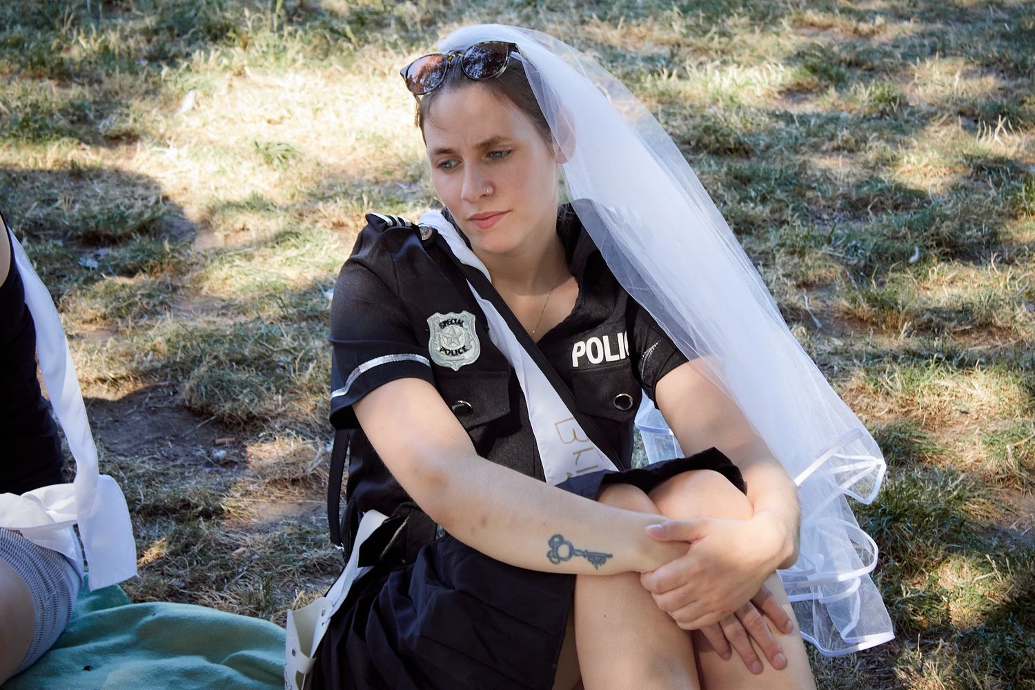 Woman wearing a police uniform shirt and bridal veil sits outdoors on grass in a creative costume combination.