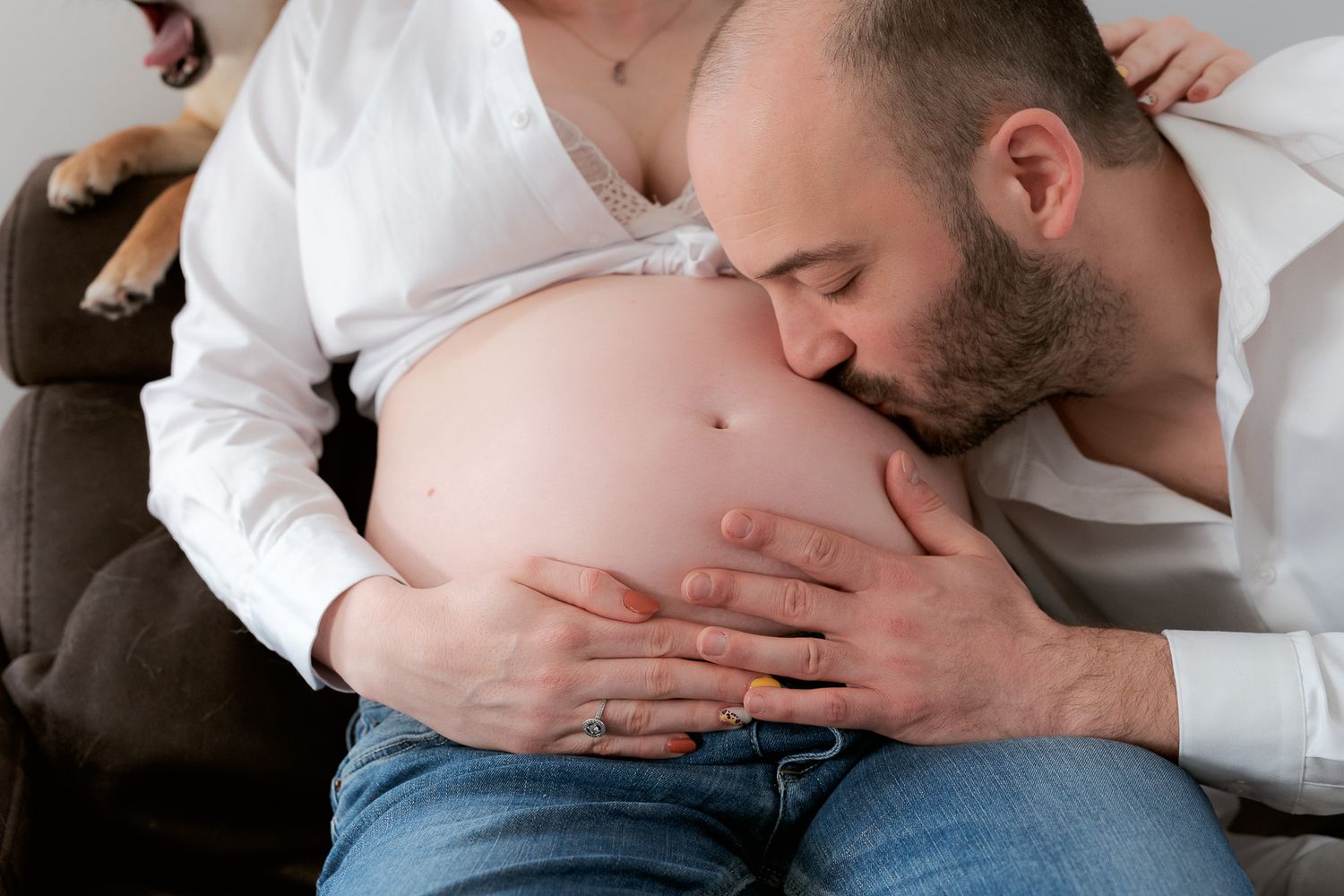 Expectant father kissing pregnant woman's bare belly while sitting together on a couch.