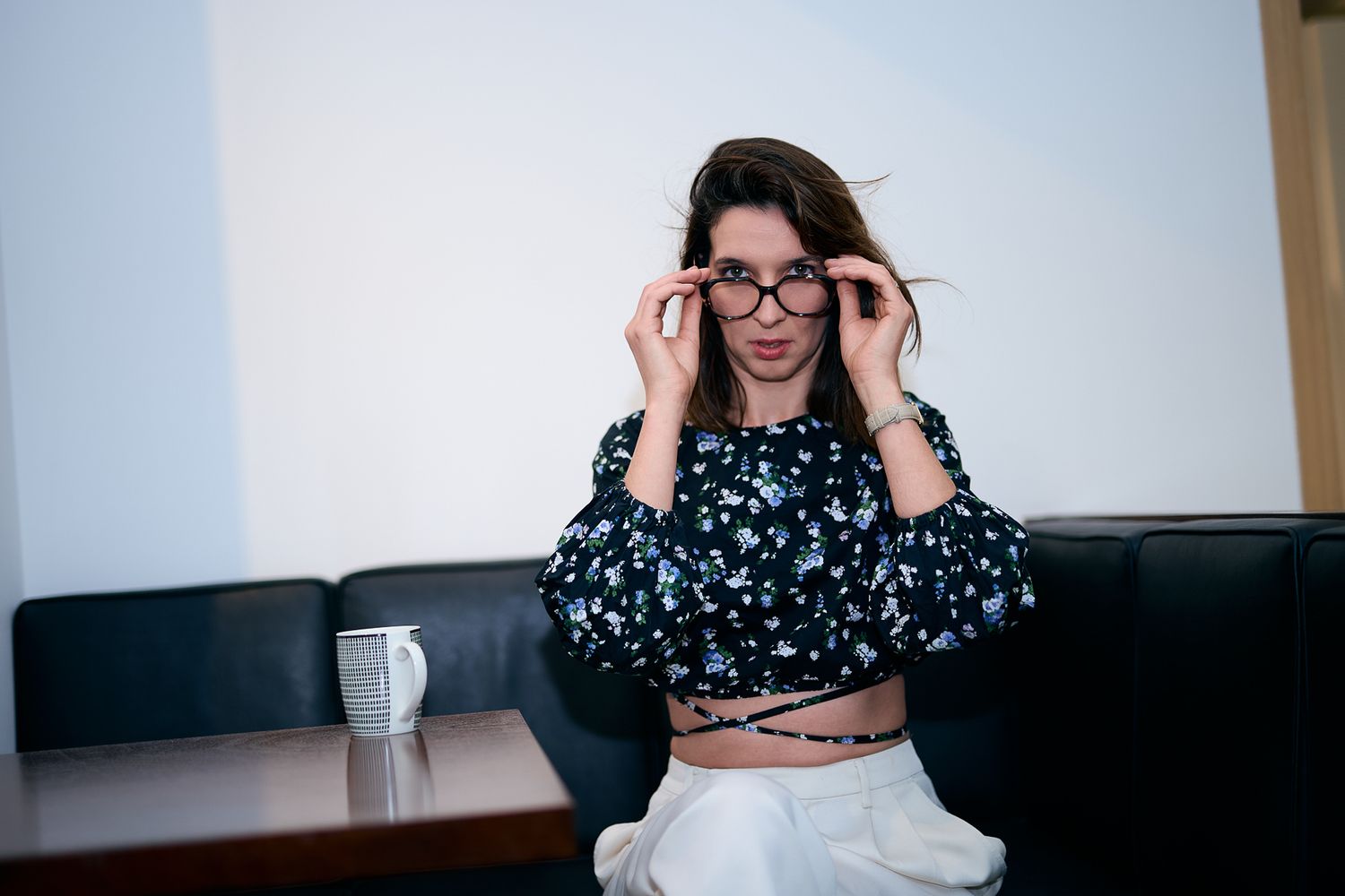 Young woman in floral crop top adjusting black glasses while sitting on dark leather sofa indoors.