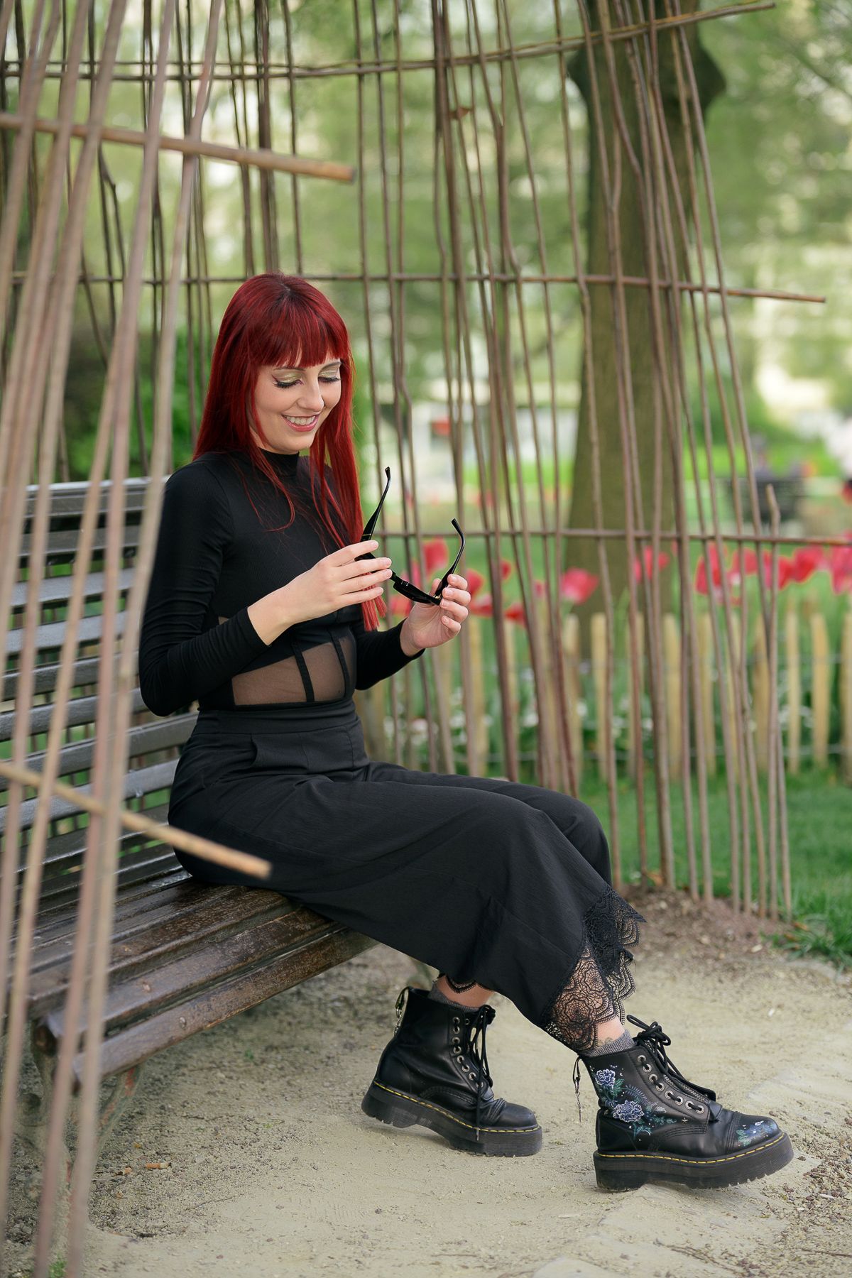 Red-haired woman in black outfit sitting on bench examining sunglasses in bamboo garden.