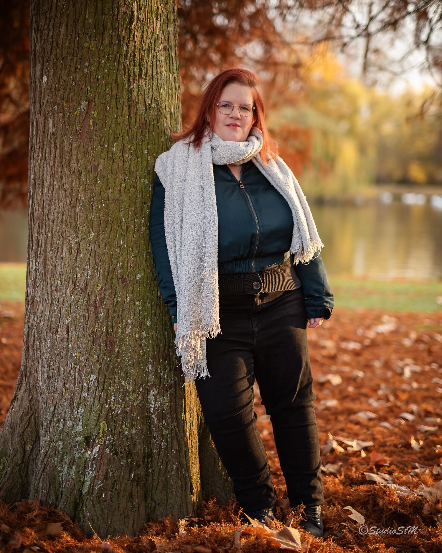 Red-haired woman in teal jacket and white scarf leaning against tree in autumn park with fallen leaves.