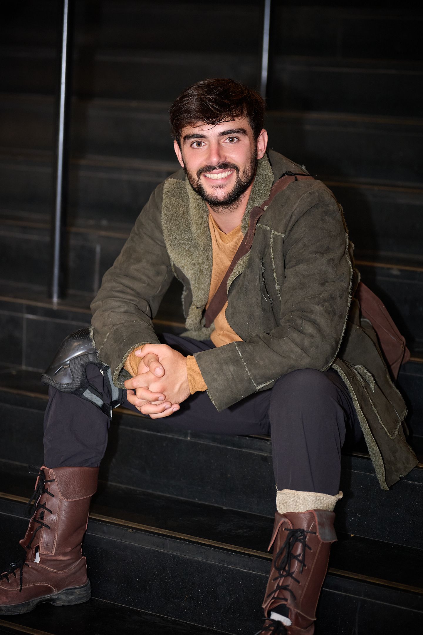 Smiling young man with beard sitting on dark steps wearing green jacket and brown lace-up boots.