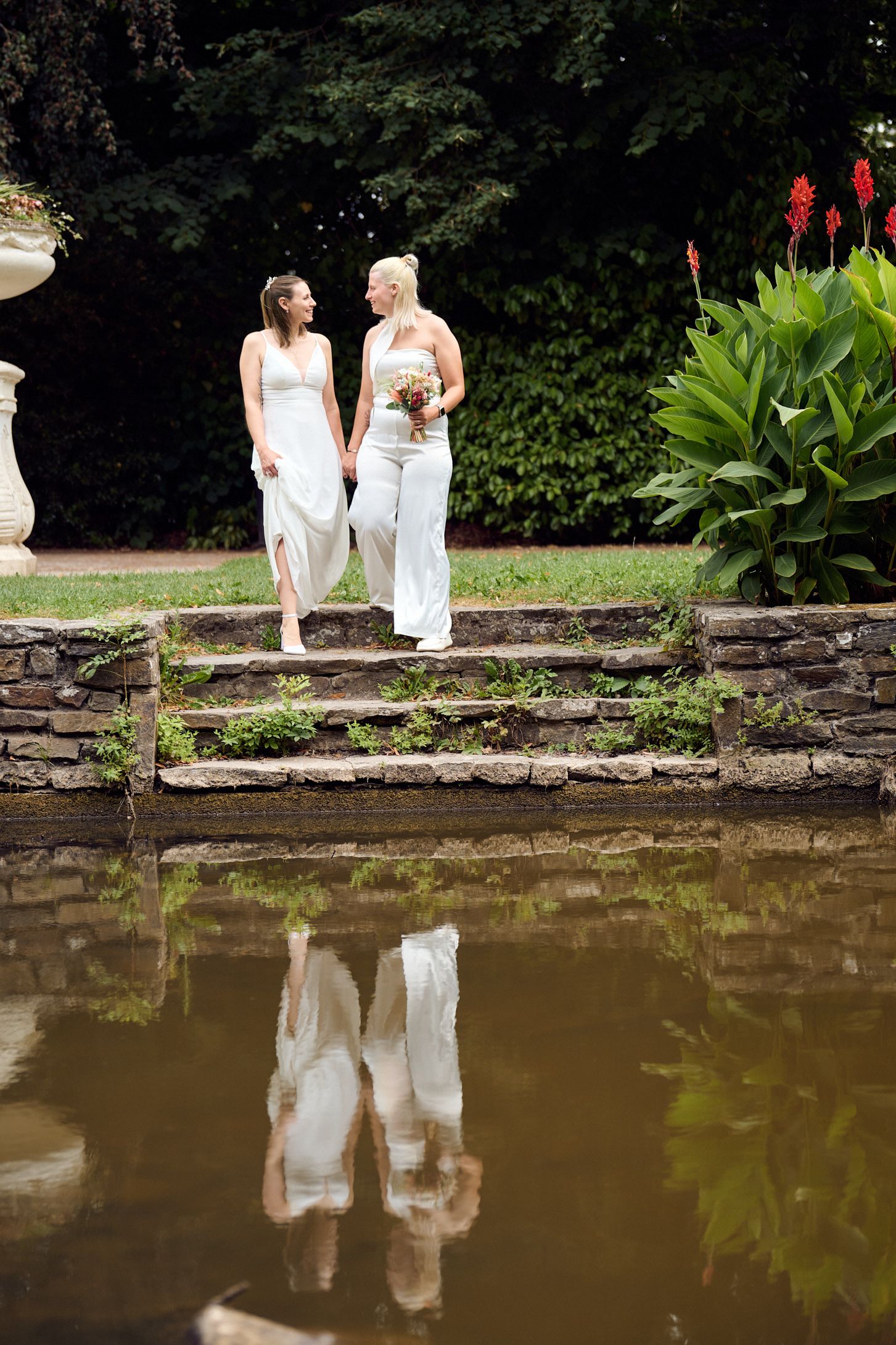Two brides in white holding flowers stand on stone steps by a pond, their reflections visible in the water.