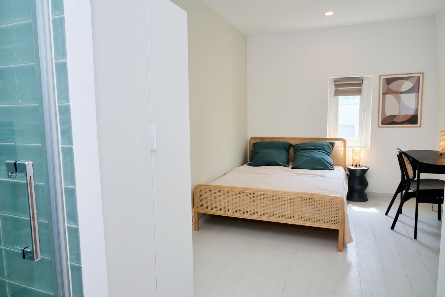Modern studio bedroom with wooden bed, teal pillows, white wardrobe, and glass shower partition on white tile floors.