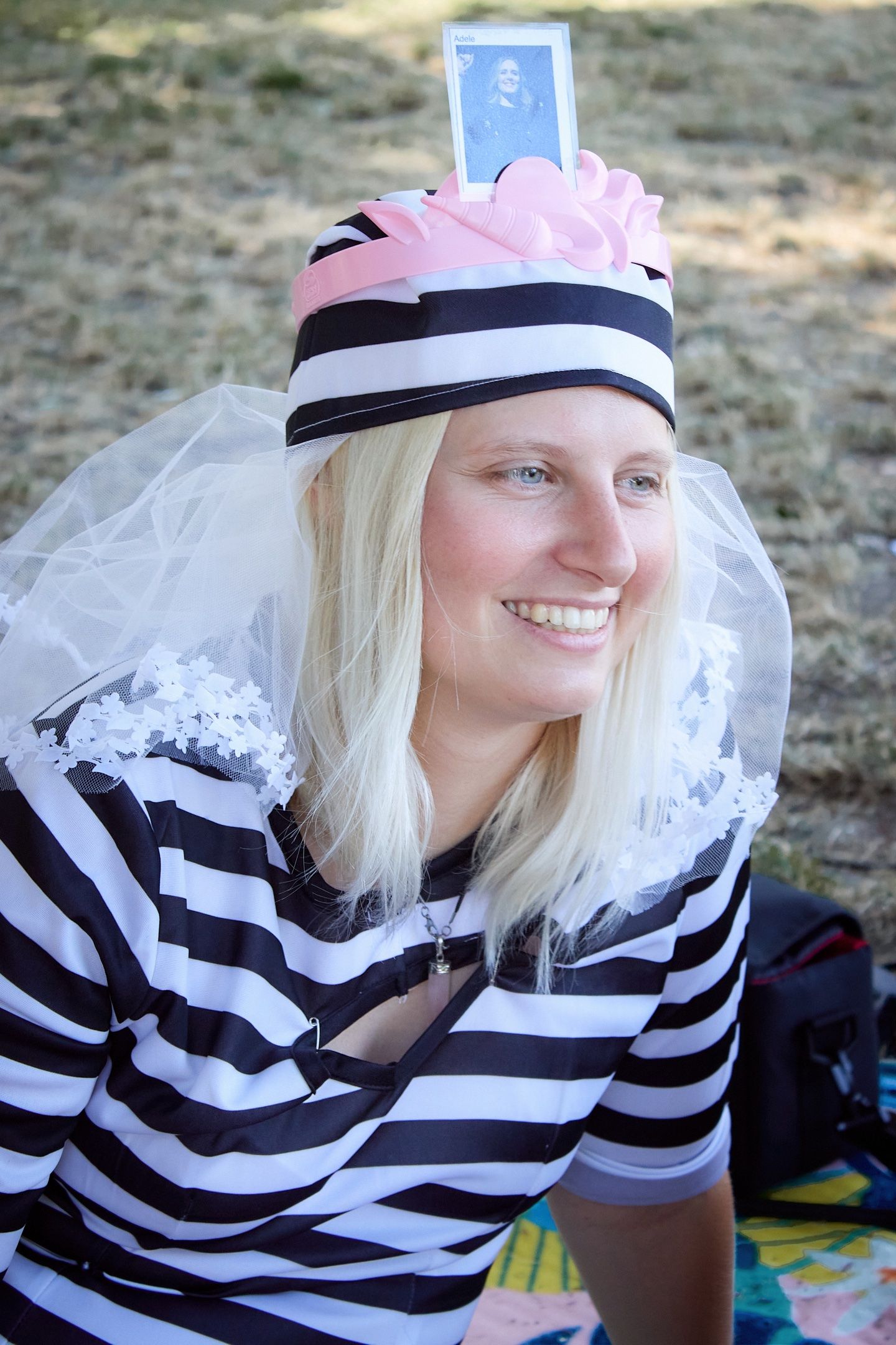 Smiling blonde woman wearing striped costume with pink bow hat, veil, and playing card at outdoor event.