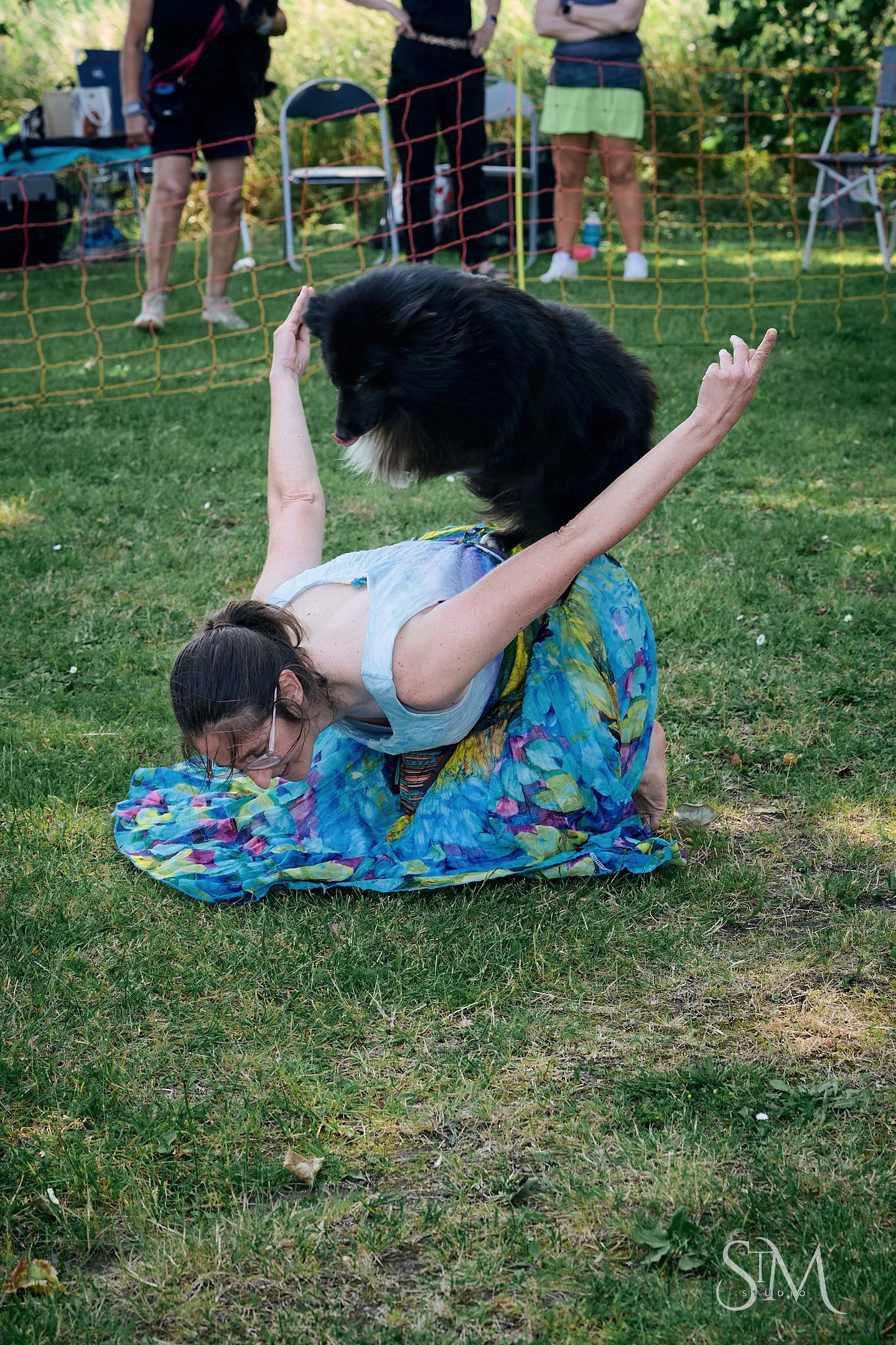 Woman in colorful skirt performs acrobatic pose on grass while a black dog jumps over her back outdoors.