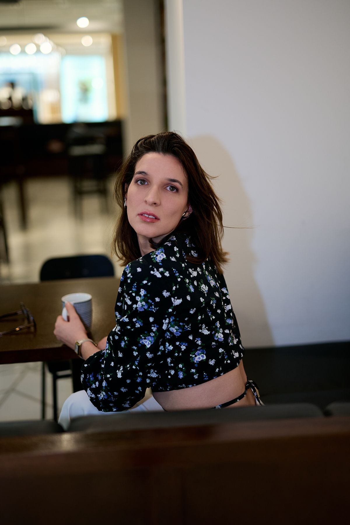 Young woman in floral black blouse holding coffee cup, sitting at cafe table, looking over shoulder.