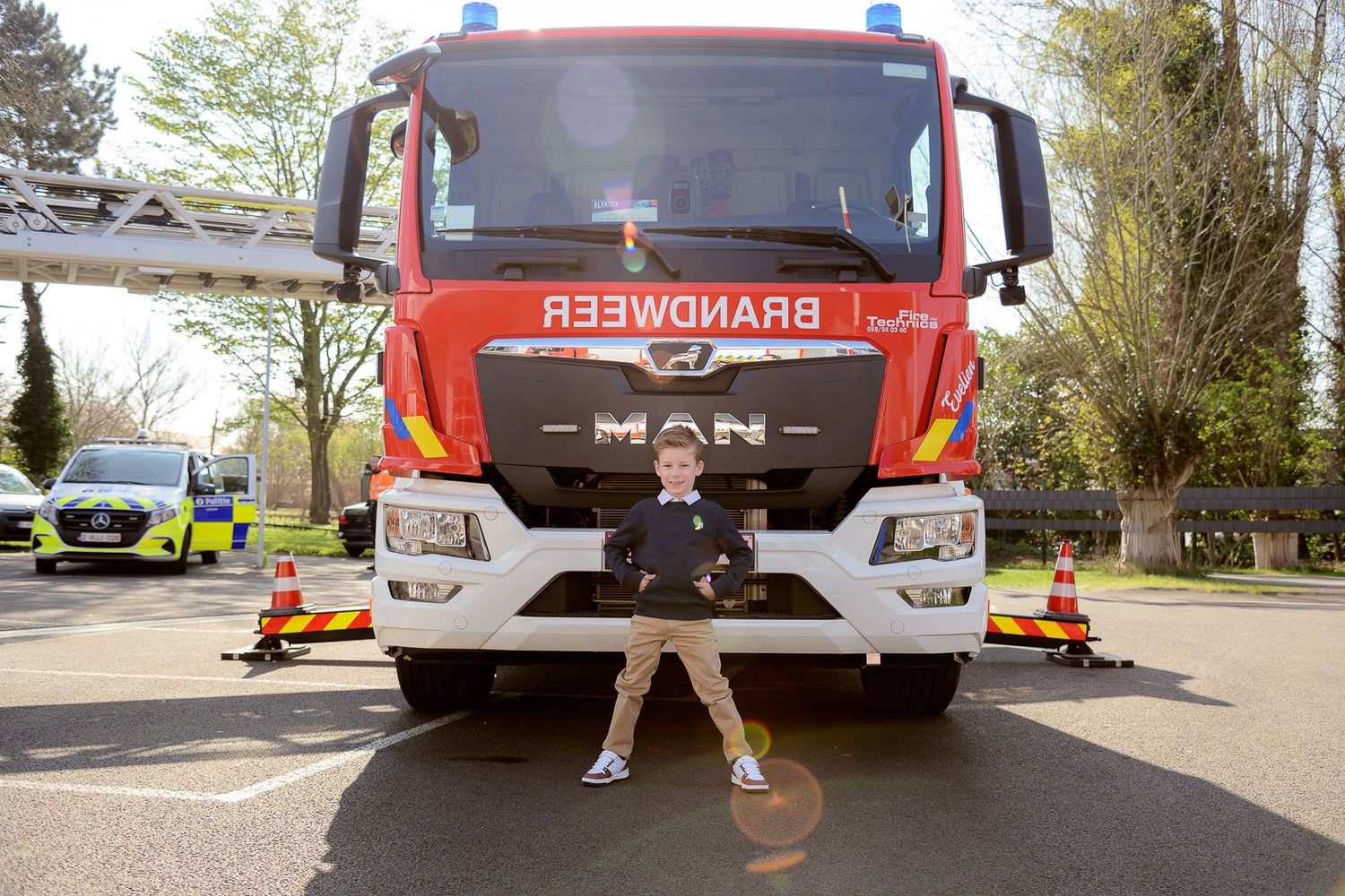 Young boy posing confidently in front of a red MAN Brandweer fire truck outdoors.