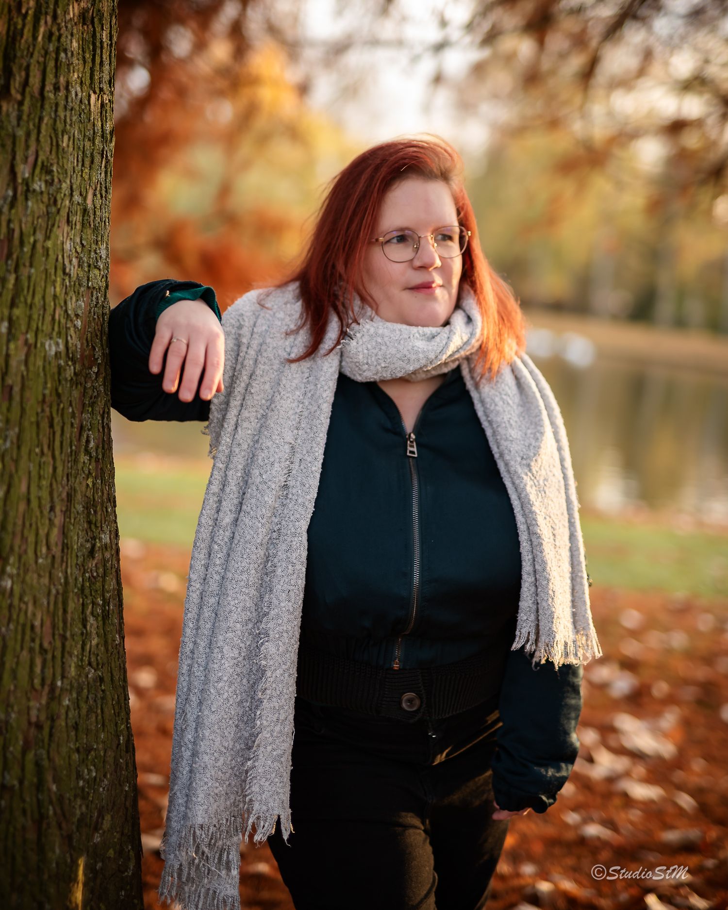 Red-haired woman in black outfit and white scarf posing by tree in colorful autumn park setting.