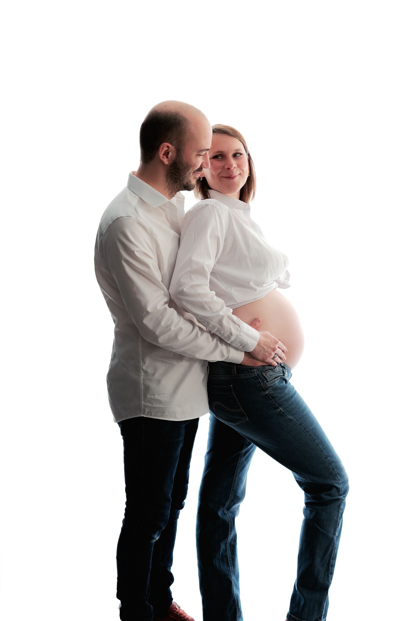 A pregnant woman in jeans and white shirt stands with her partner, who embraces her from behind on white background.