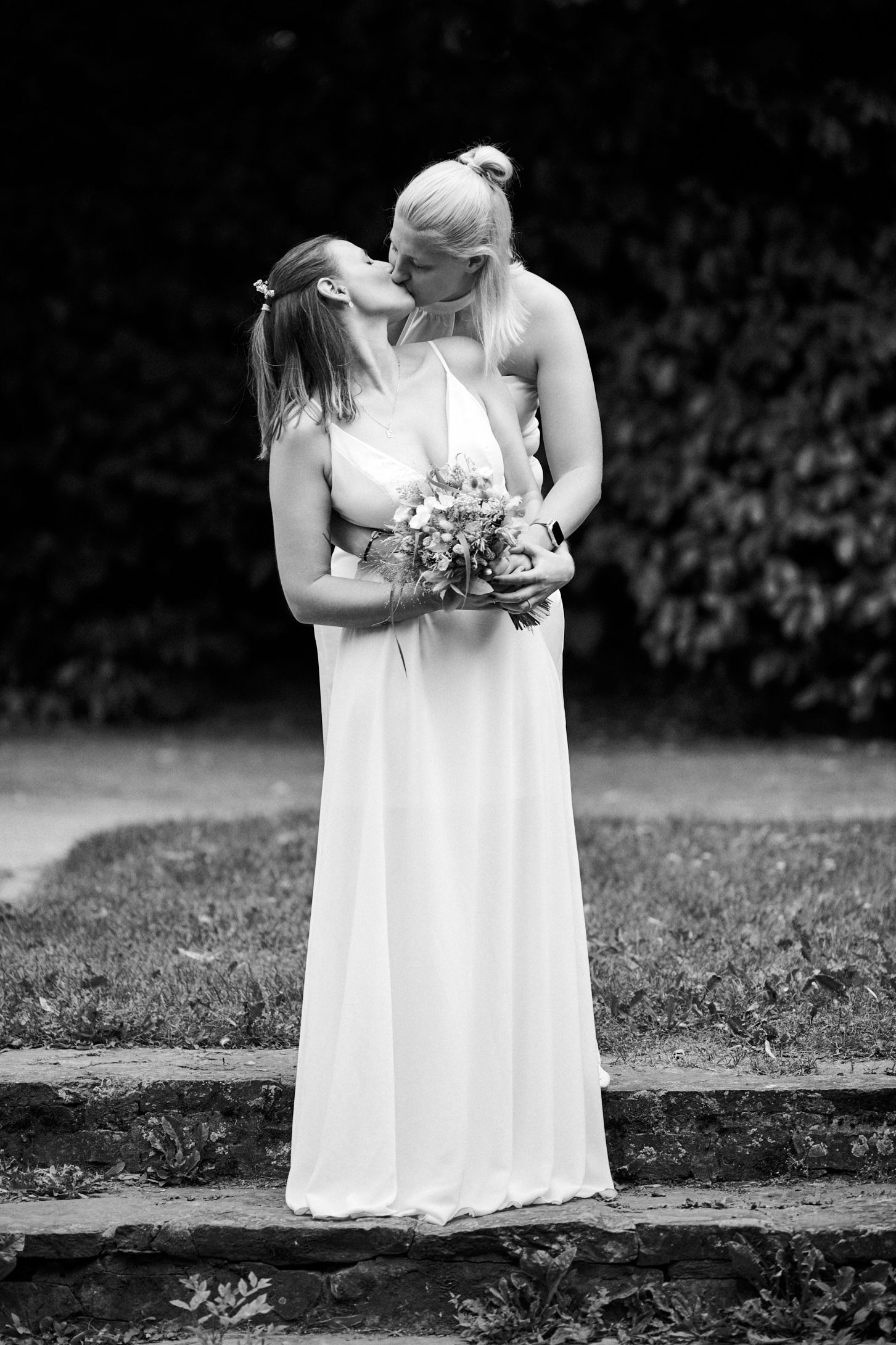 Two brides sharing a kiss in a garden, one holding a bouquet, wearing elegant white gowns in a black and white photo.