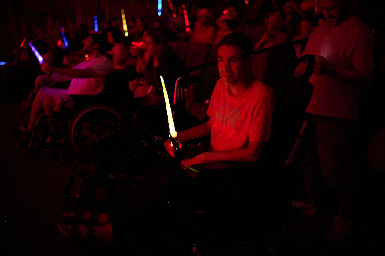 Person in wheelchair holding glowing green light stick at a dark concert venue with colorful lights in background.