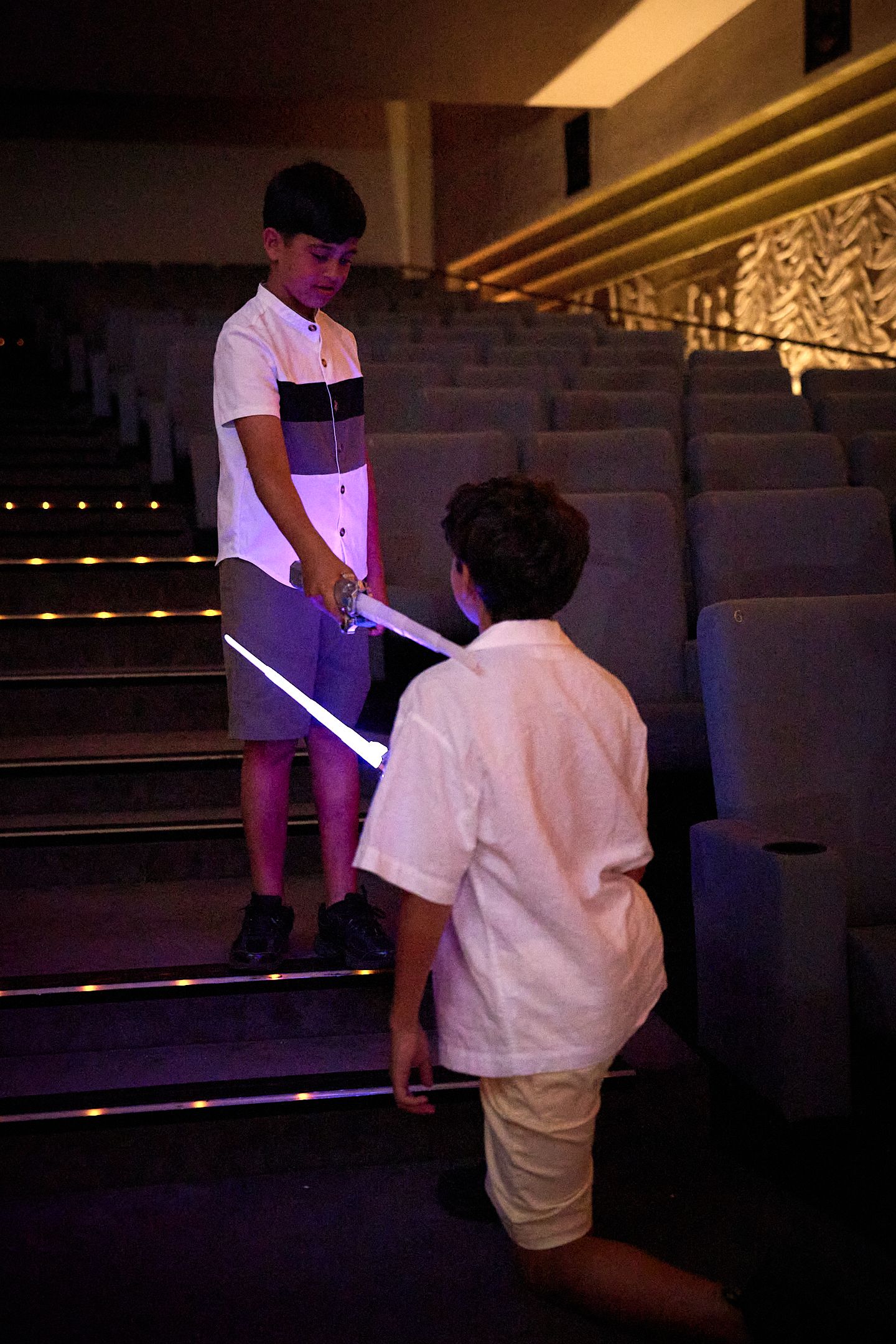 Two boys playing with light-up toy lightsabers on theater steps in a dimly lit auditorium.