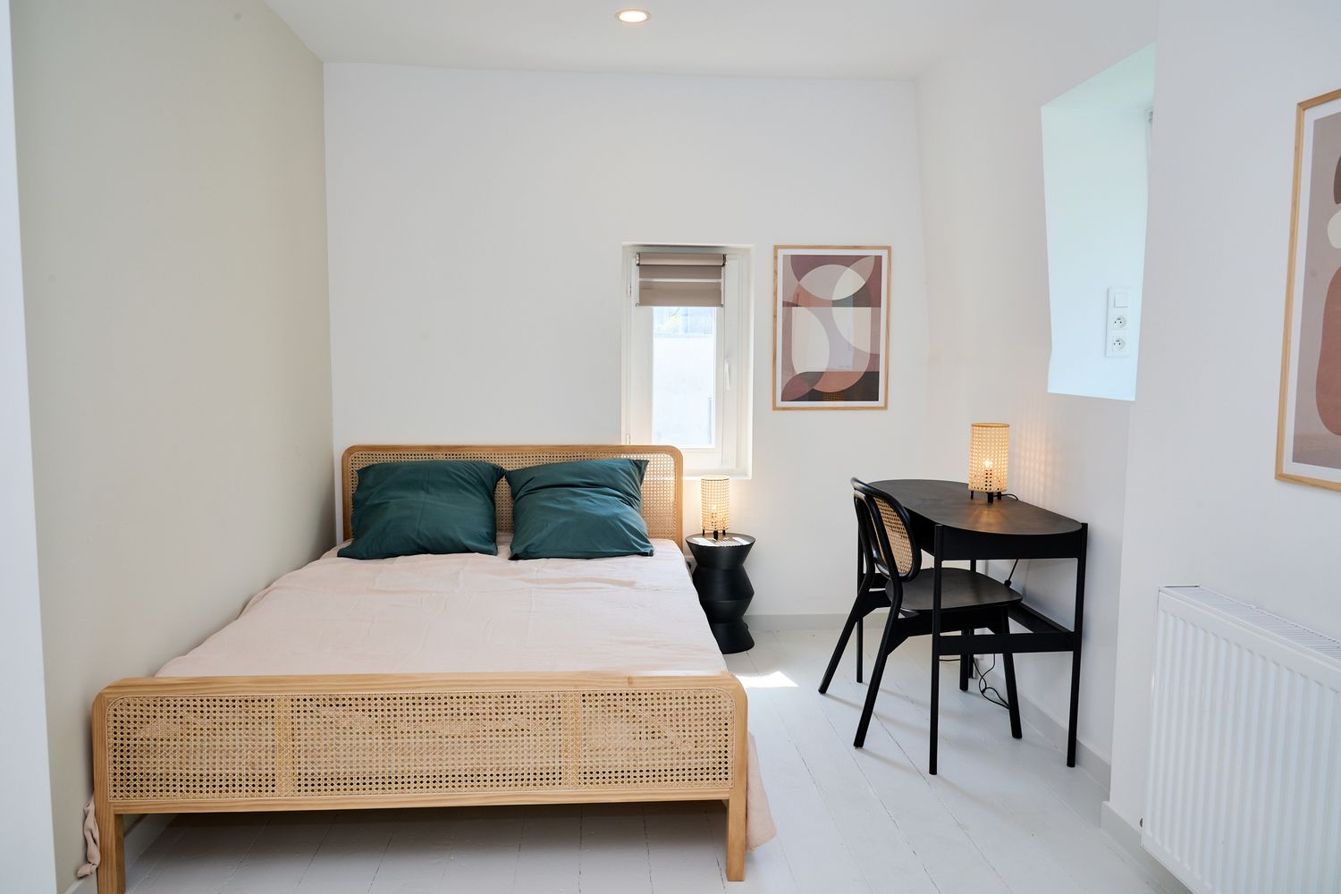 Minimalist white bedroom with rattan bed, teal pillows, black desk and chair, and framed wall art.