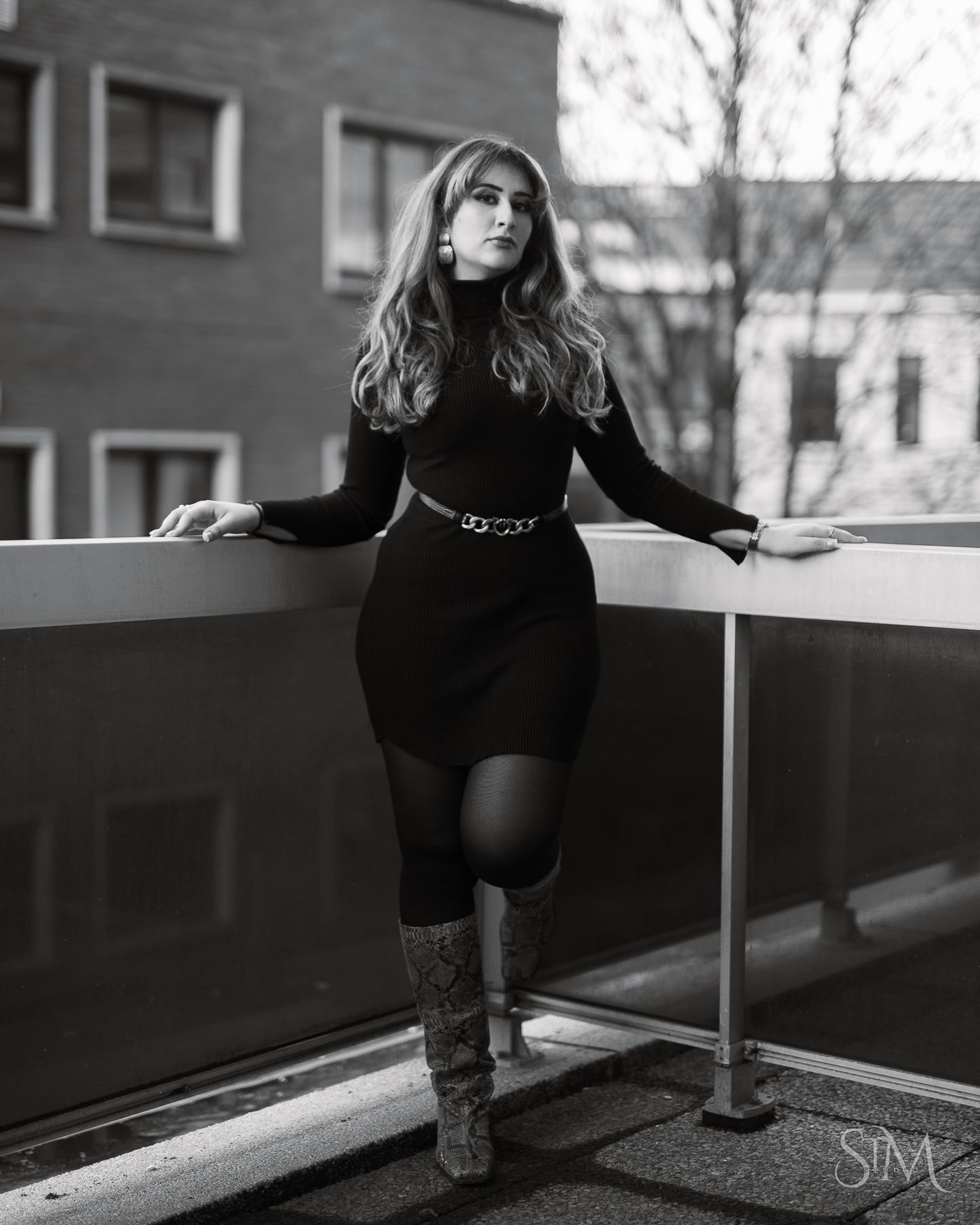 Black and white photo of a woman in a black turtleneck dress and boots posing on a balcony.