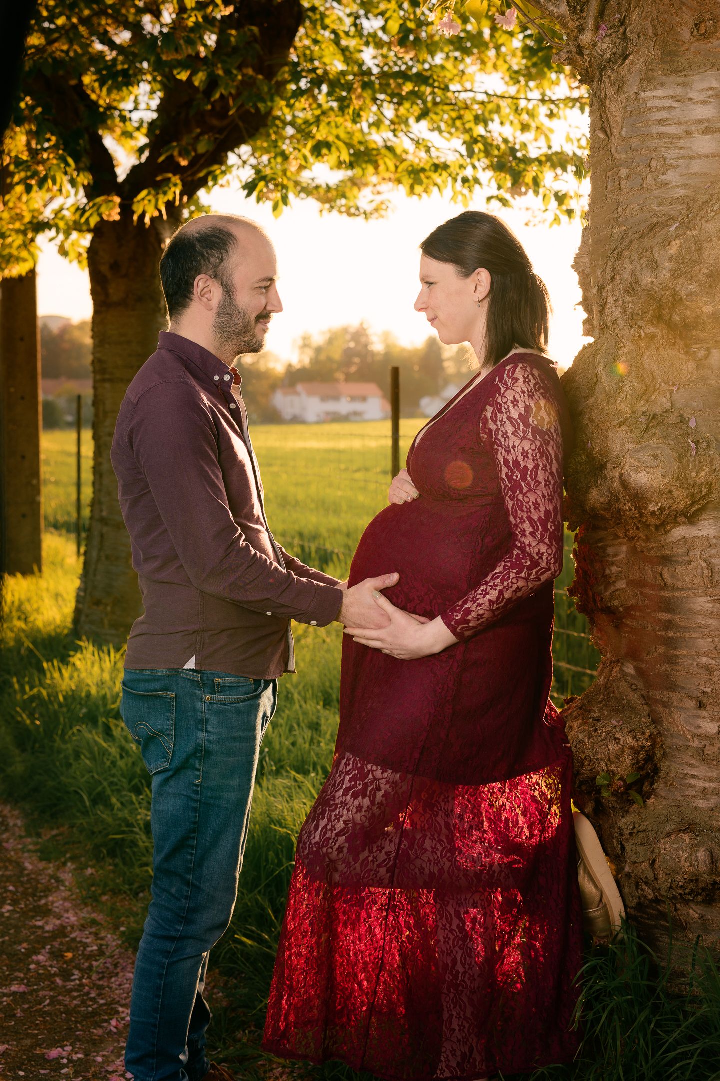 Couple sharing a tender moment by a tree at golden hour, pregnant woman in red lace dress cradling her baby bump.