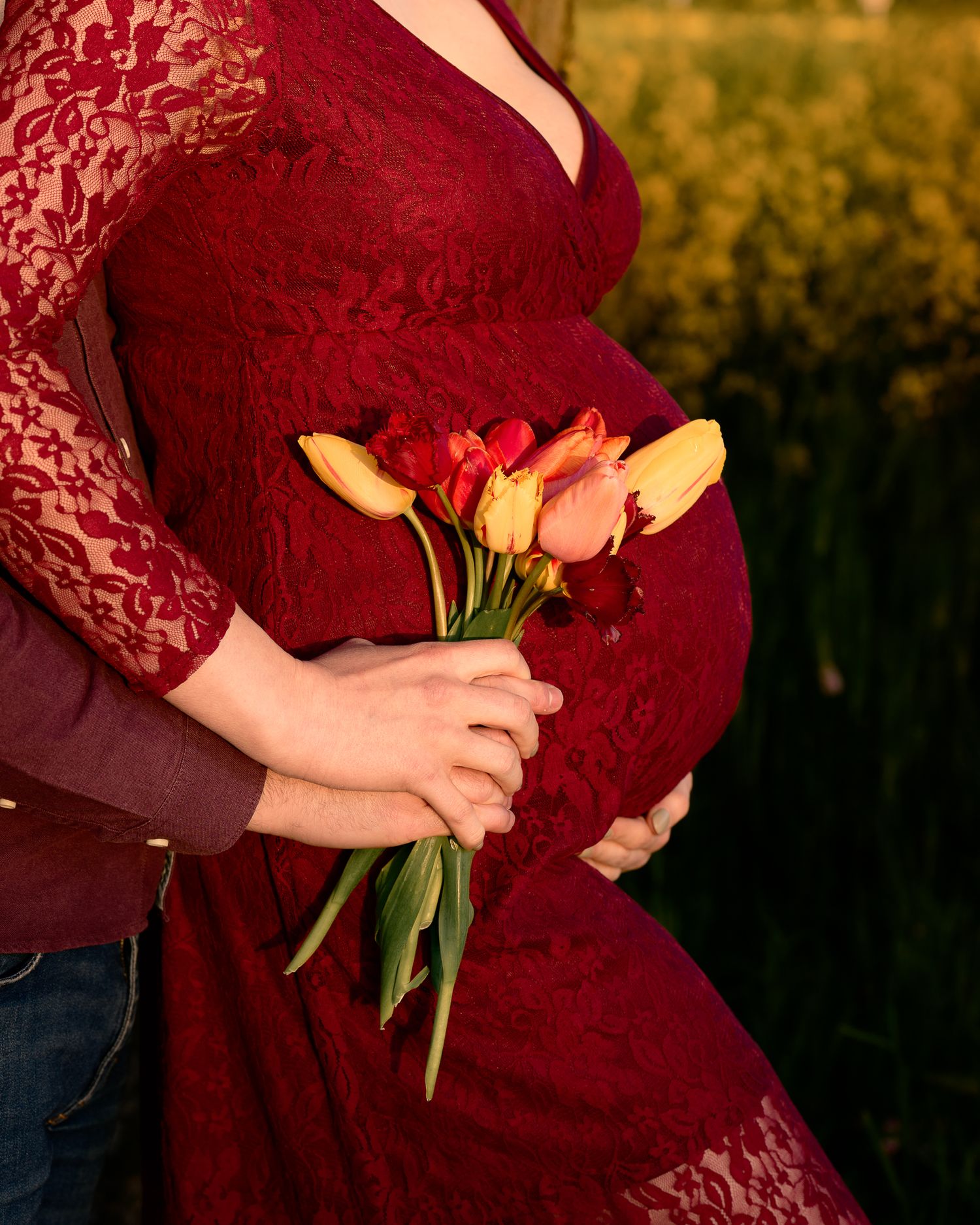 Pregnant woman in red lace dress holds colorful tulips with partner outdoors in golden field.