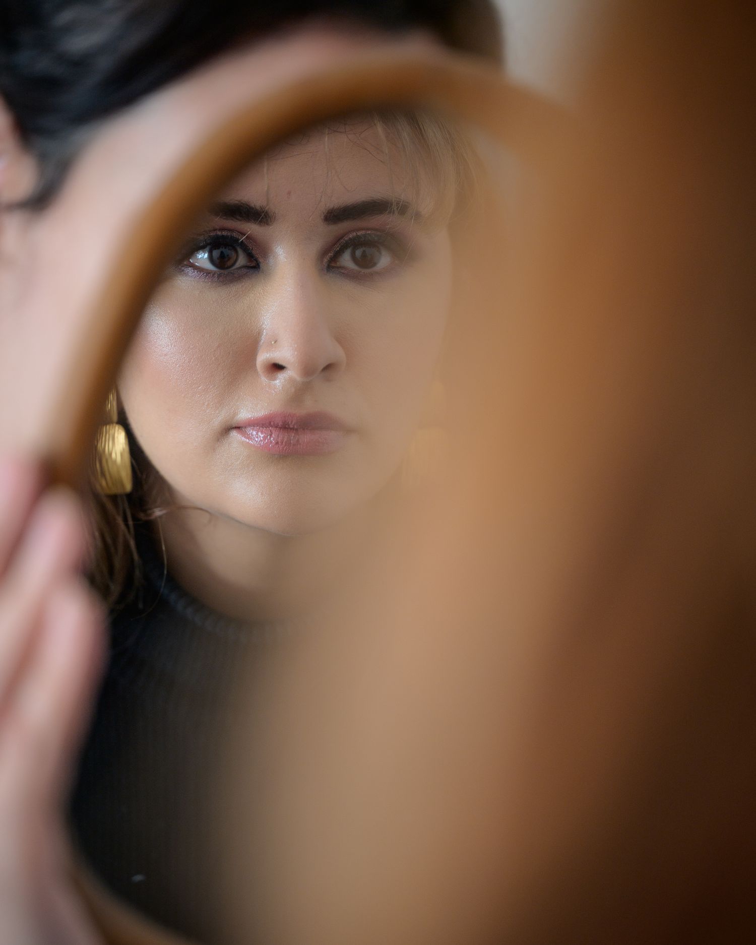 Woman with gold earrings gazes thoughtfully at her reflection in a handheld mirror.