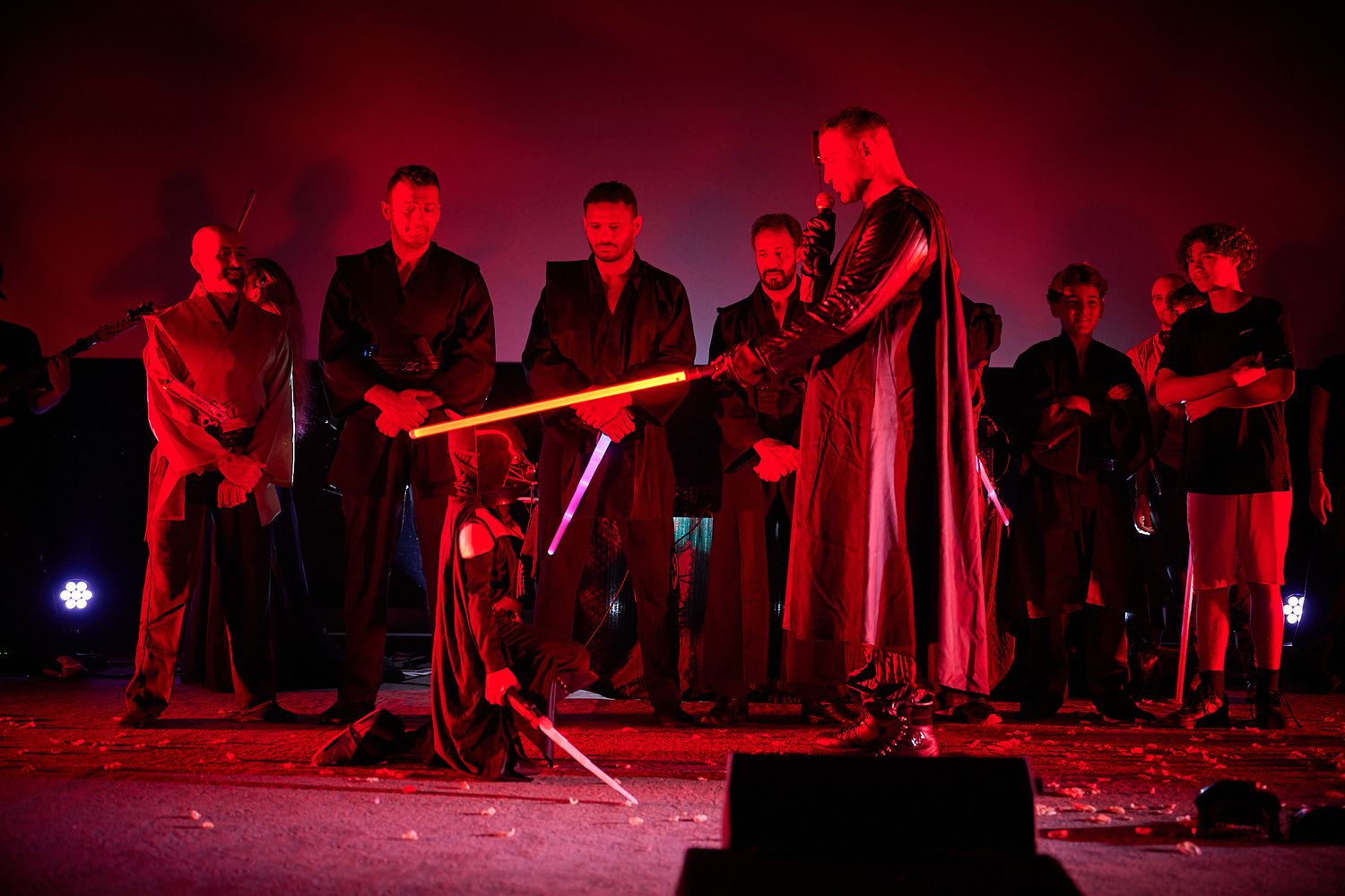 Group of performers in dark cloaks on stage with glowing red lightsabers during a dramatic theatrical event.