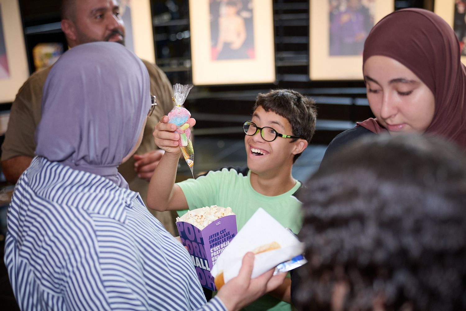 A smiling boy in glasses shares popcorn with friends at a casual gathering, surrounded by people in hijabs.