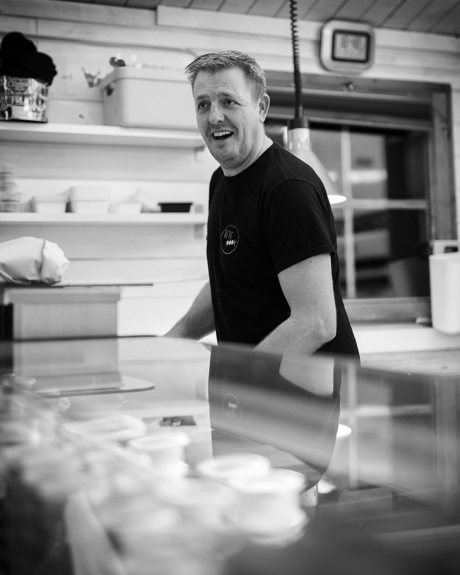 Smiling man in black t-shirt working behind a deli counter in a black and white kitchen photo.
