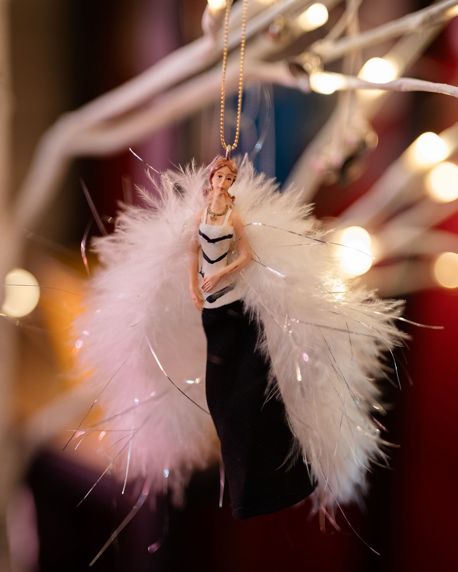 Elegant angel Christmas ornament with white feather wings and black gown, hanging on a gold chain with bokeh lights.