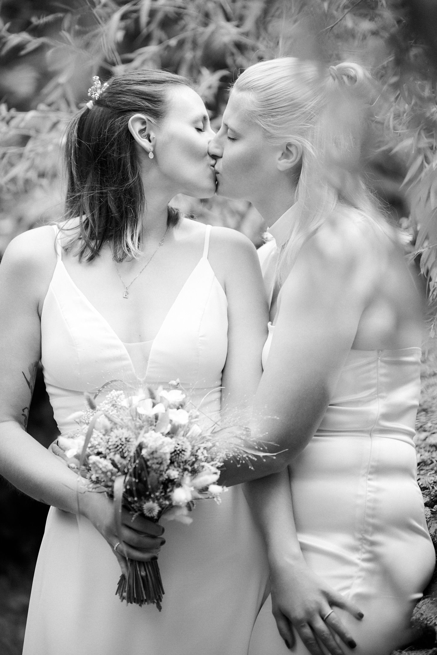 Two brides in white dresses share a kiss while holding a floral bouquet in a black and white wedding photo.