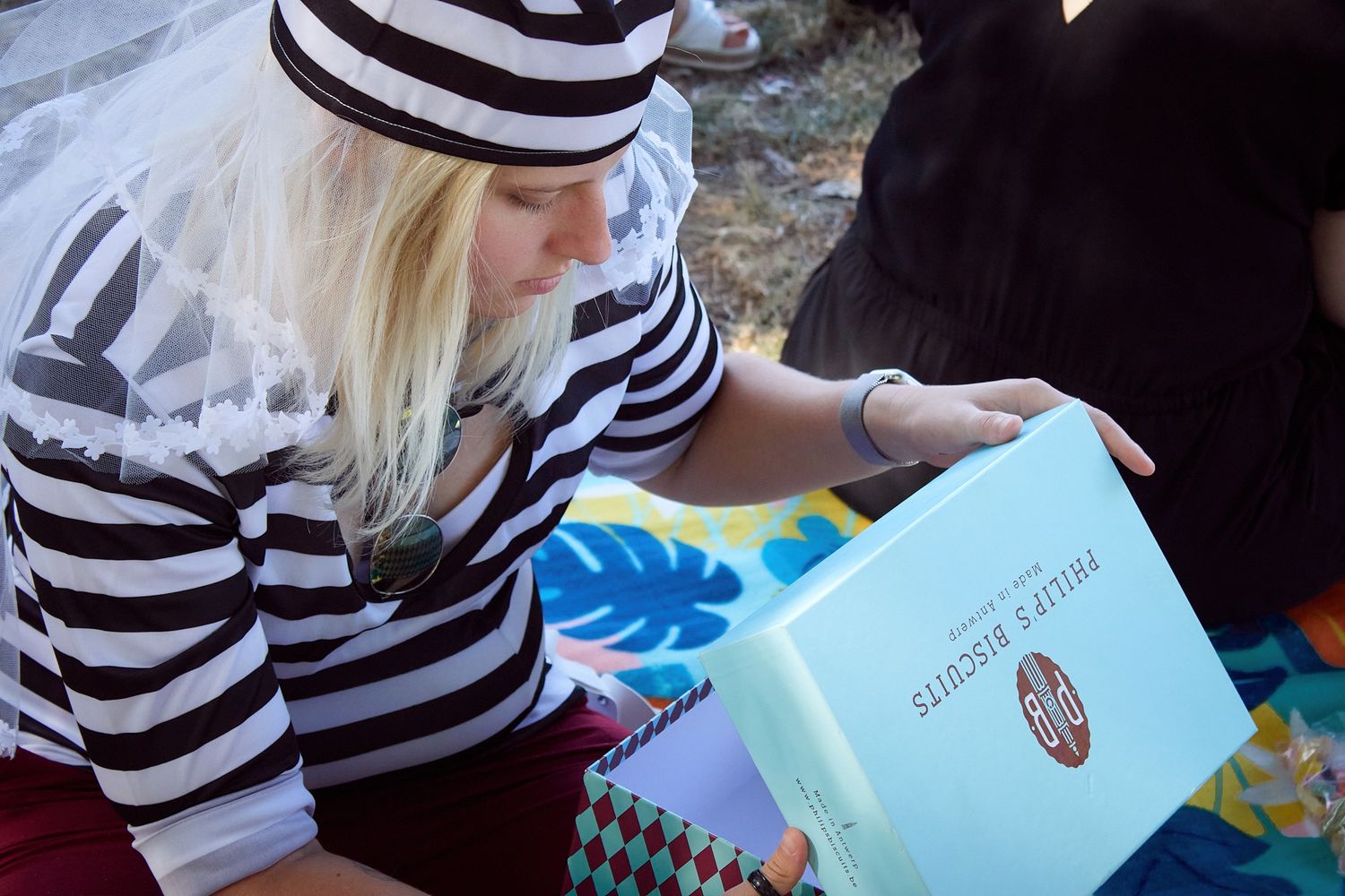 Blonde woman in striped shirt reading a light blue book outdoors at a casual gathering.