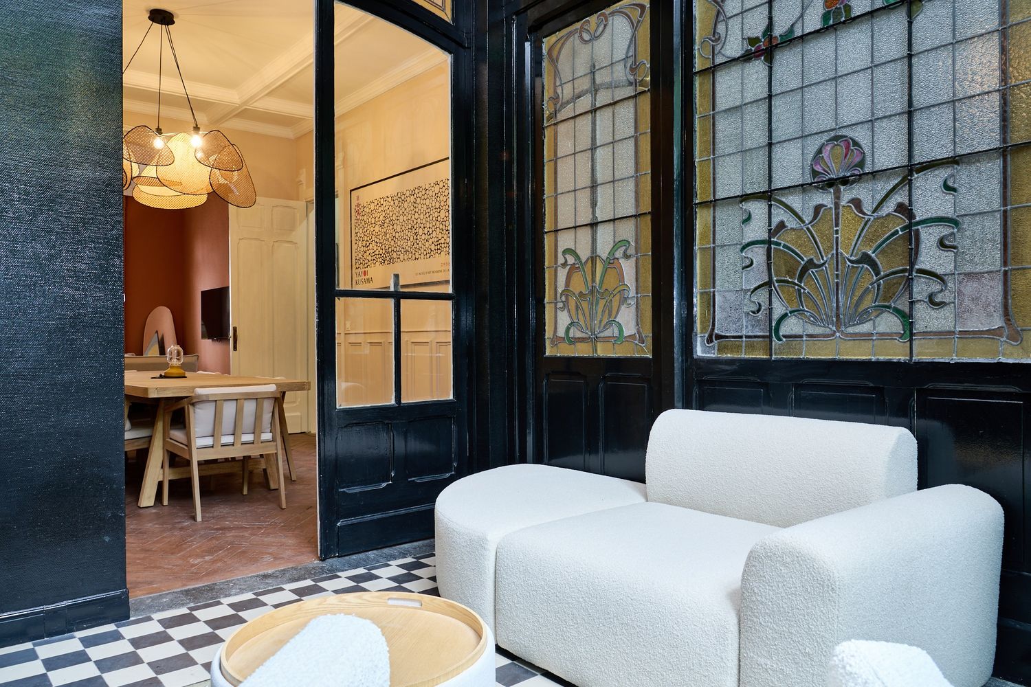 Elegant interior with white armchairs, stained glass windows, and black-framed doors opening to a dining room with checkered floors.