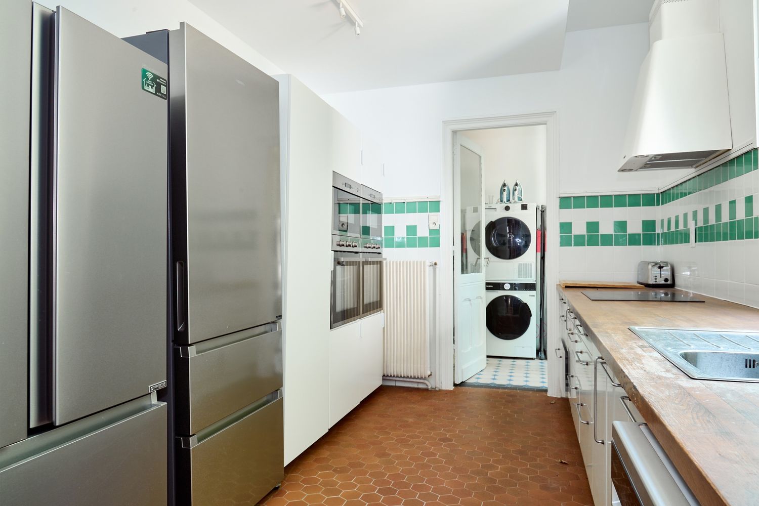 Modern kitchen with stainless steel fridge, built-in oven, stacked washer-dryer, green tile accents, and wooden countertops.