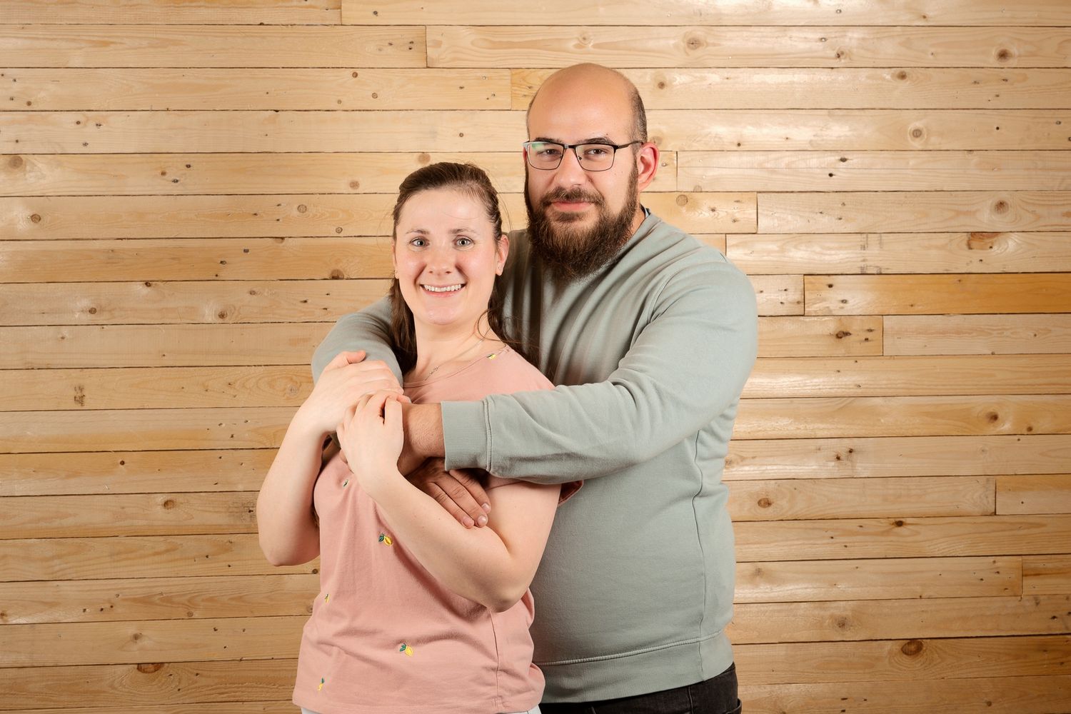 A bald man with glasses and beard hugs a smiling woman in a pink top against a wooden wall backdrop.