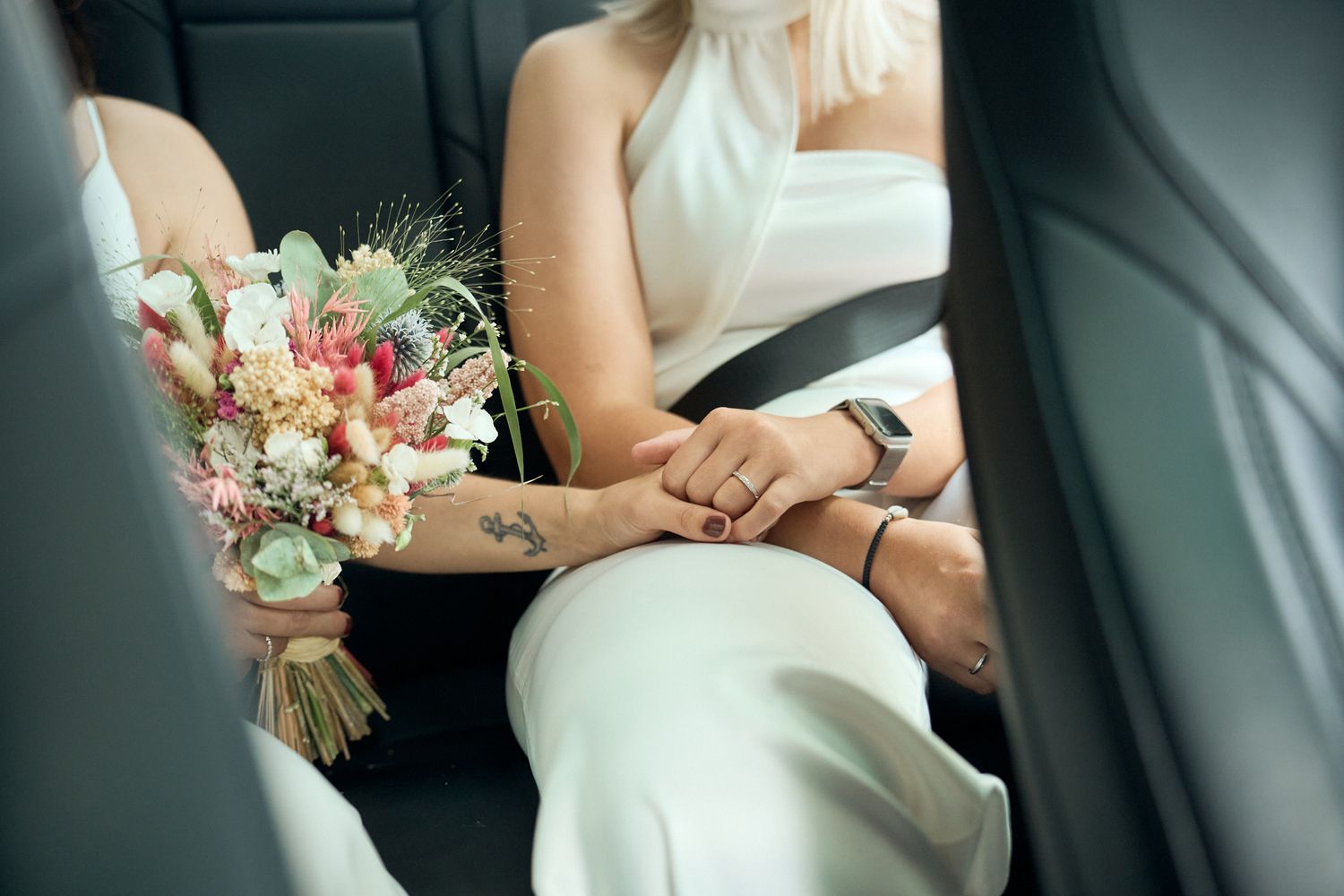 Two brides in white dresses hold hands in a car, one holding a pink and white wedding bouquet.