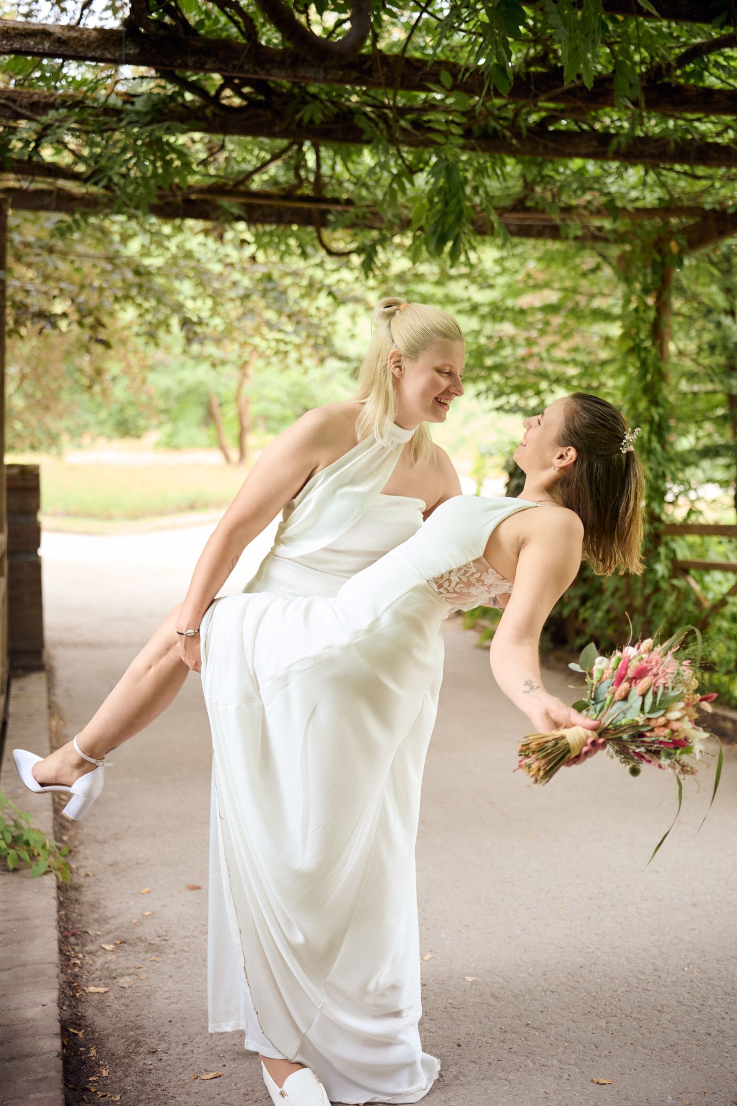 Two brides in white gowns share a joyful moment, one lifting the other, holding a colorful bouquet in a lush garden.