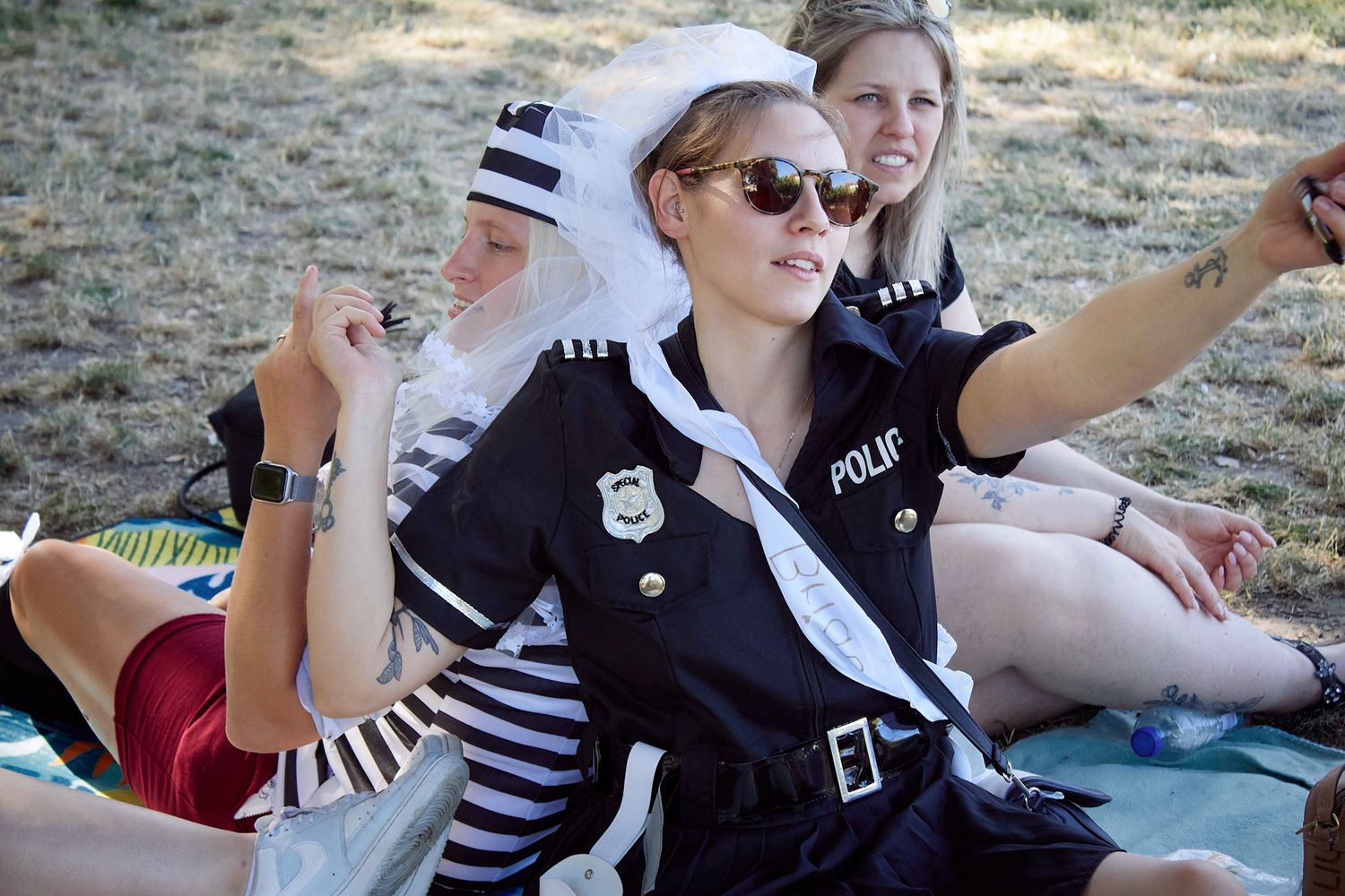 Women in costume outfits, one dressed as a police officer with bridal veil, sitting outdoors at a sunny event.