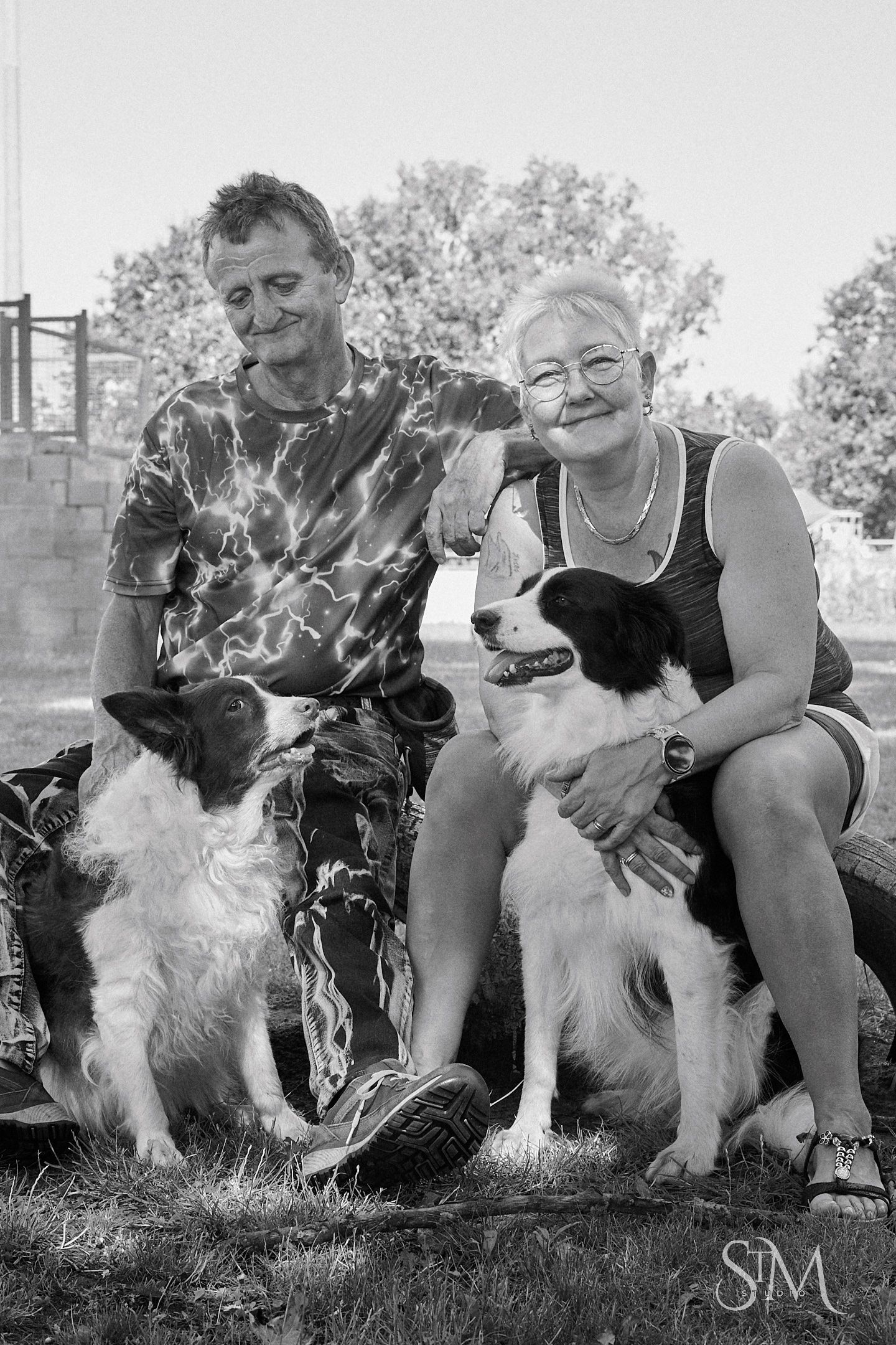 Black and white photo of smiling couple sitting outdoors with two border collie dogs on grass.
